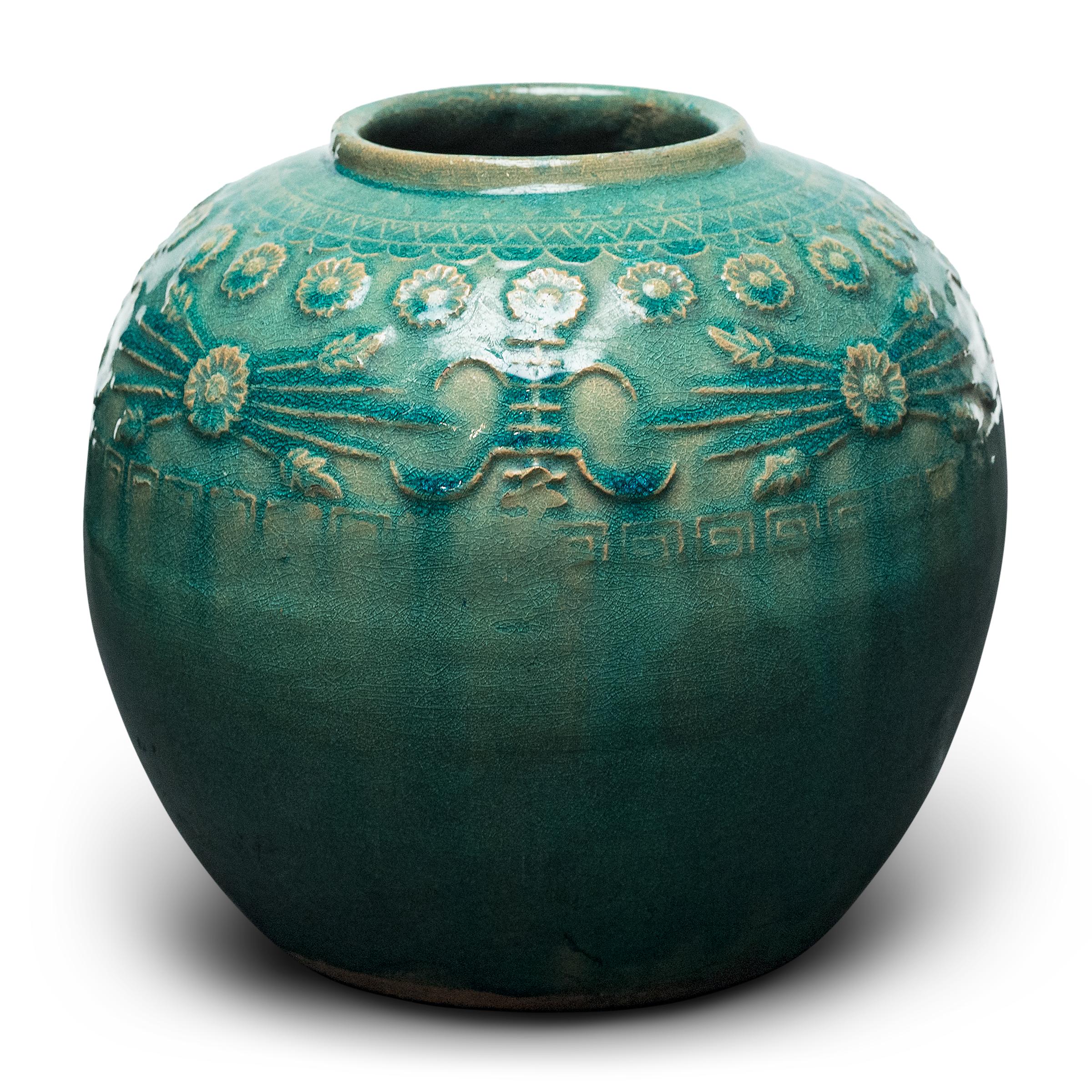 A glassy, blue-green glaze sheets, pools, and drips across the rounded body of this provincial kitchen pot, lingering on the low relief decoration to the jar's shoulders. Among the raised patterns are round chrysanthemum blossoms and stylized Shou