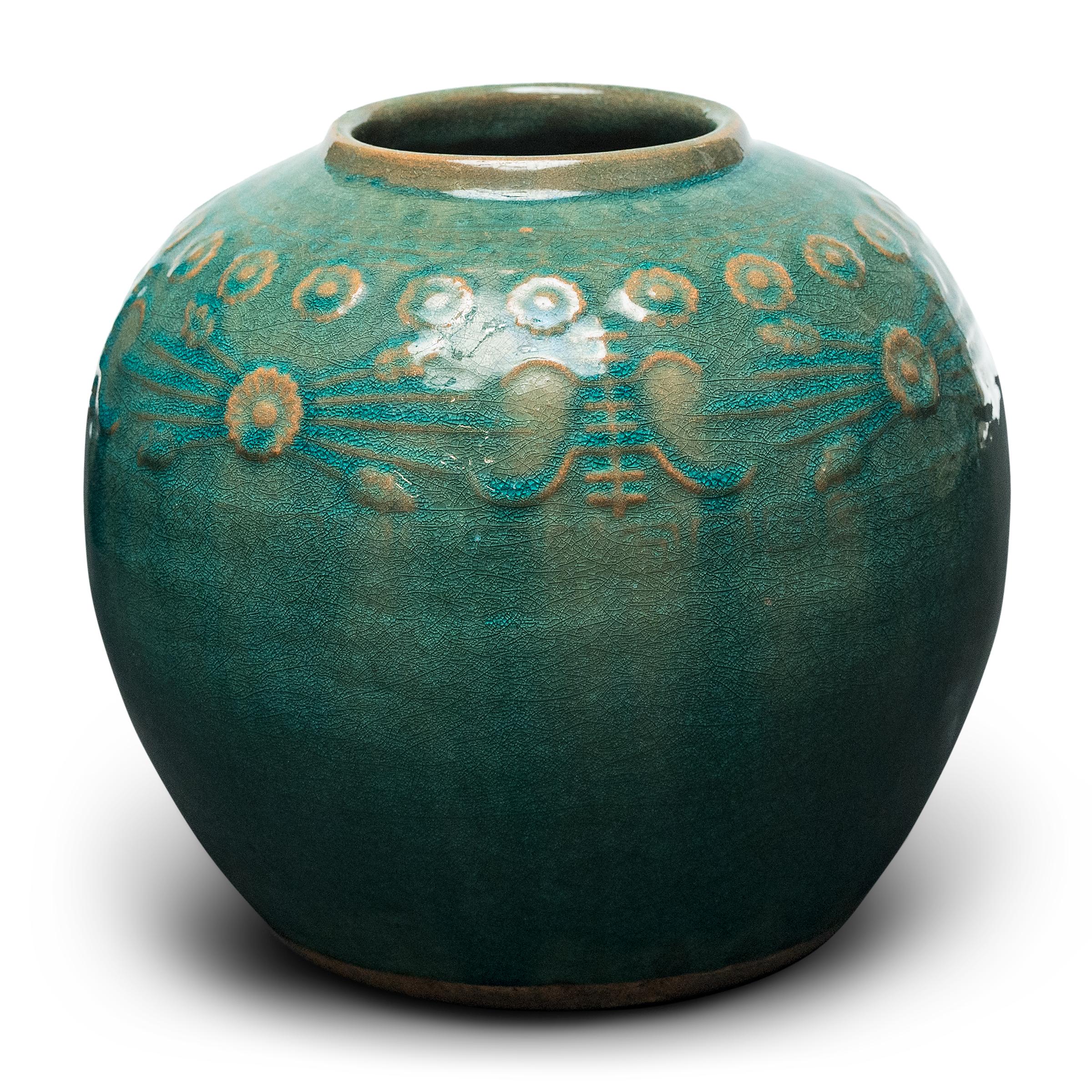 A glassy, blue-green glaze sheets, pools, and drips across the rounded body of this provincial kitchen pot, lingering on the low relief decoration to the jar's shoulders. Among the raised patterns are round chrysanthemum blossoms and stylized Shou