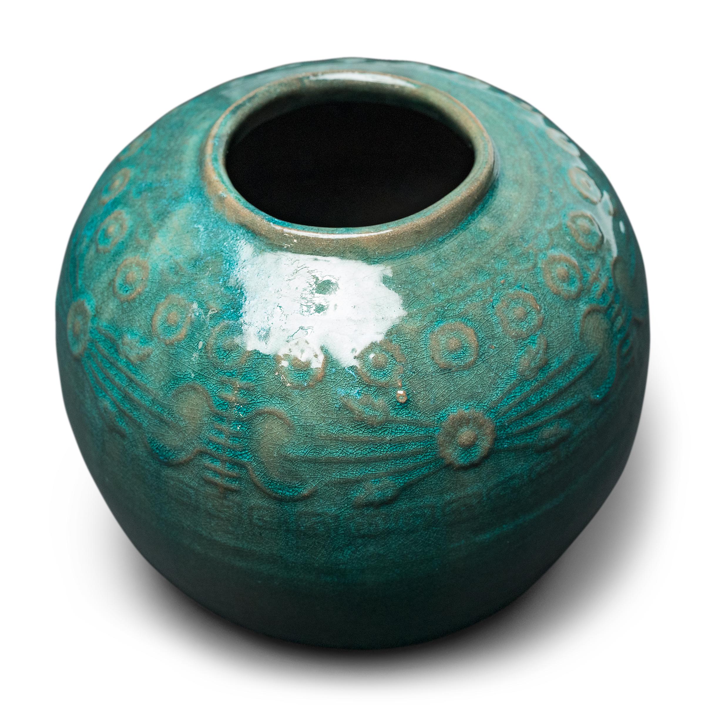 A glassy, blue-green glaze sheets, pools, and drips across the rounded body of this provincial kitchen pot, lingering on the low relief decoration to the jar's shoulders. Among the raised patterns are round chrysanthemum blossoms and stylized Shou