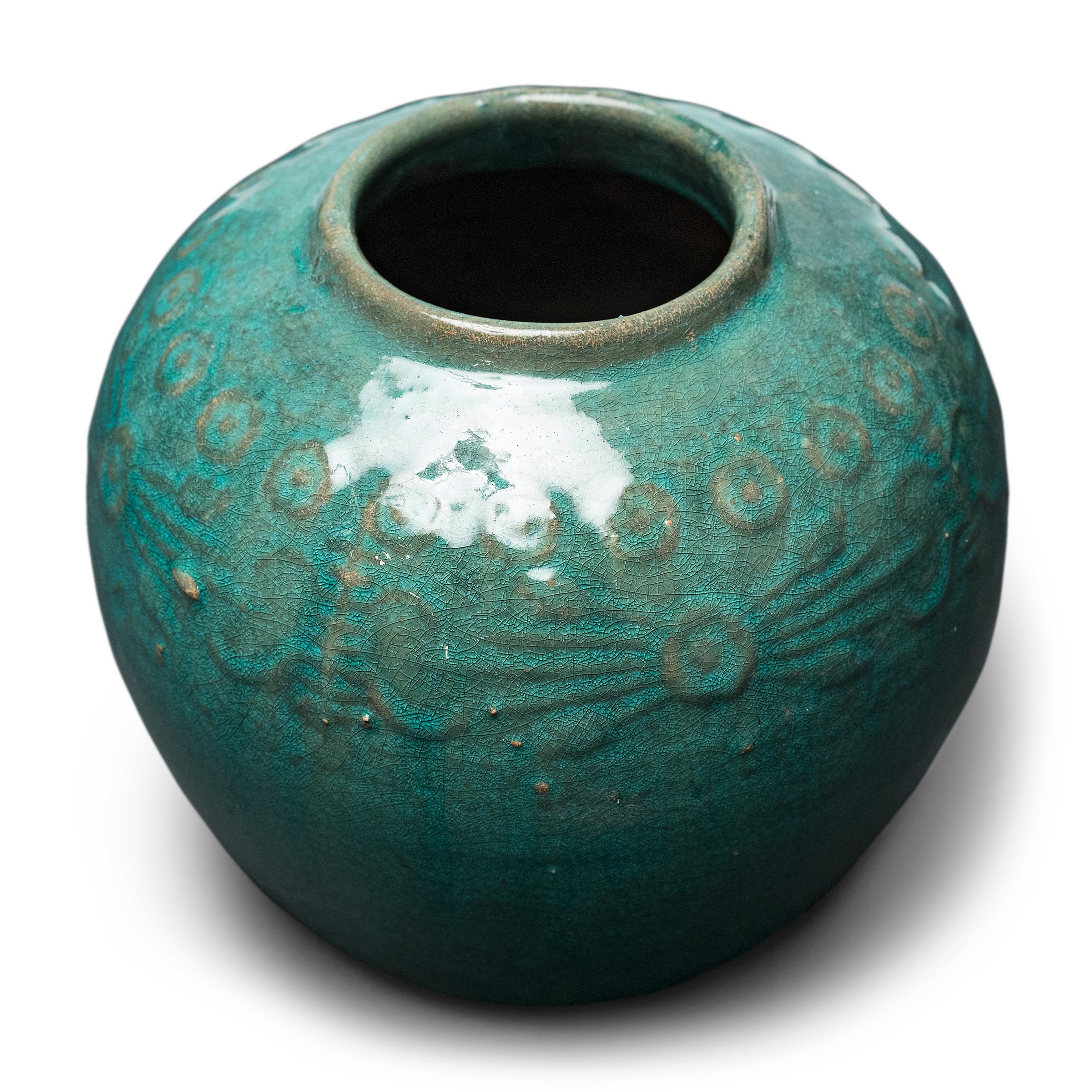 A glassy, blue-green glaze sheets, pools, and drips across the rounded body of this provincial kitchen pot, lingering on the low relief decoration to the jar's shoulders. Among the raised patterns are round chrysanthemum blossoms and stylized Shou