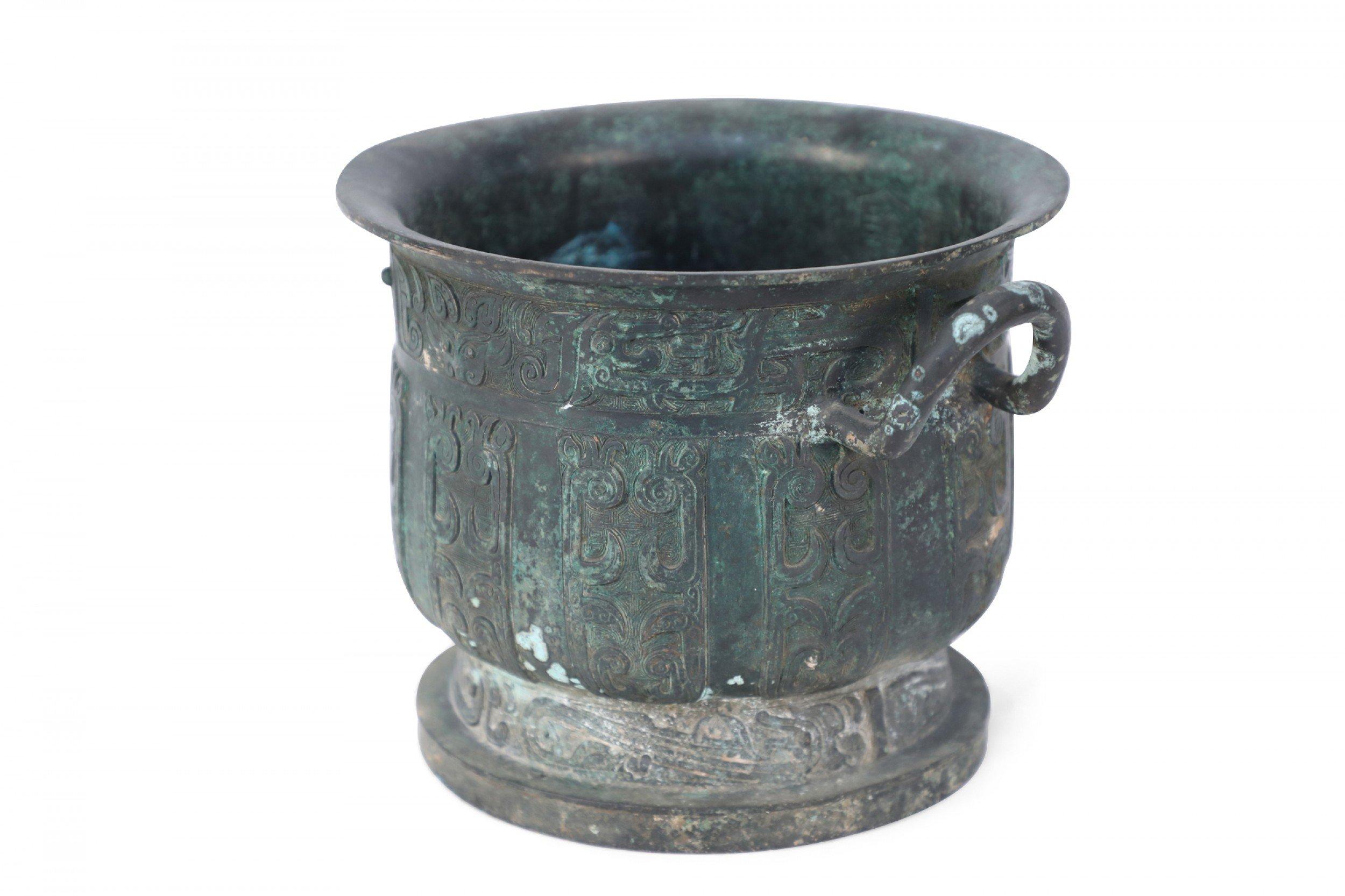 Antica copia in stile Dinastia Han cinese di un'urna in bronzo patinato scolpita con bande geometriche in rilievo che ne avvolgono la forma, seduta in cima a un piede rotondo e dotata di due manici curvi e verticali.