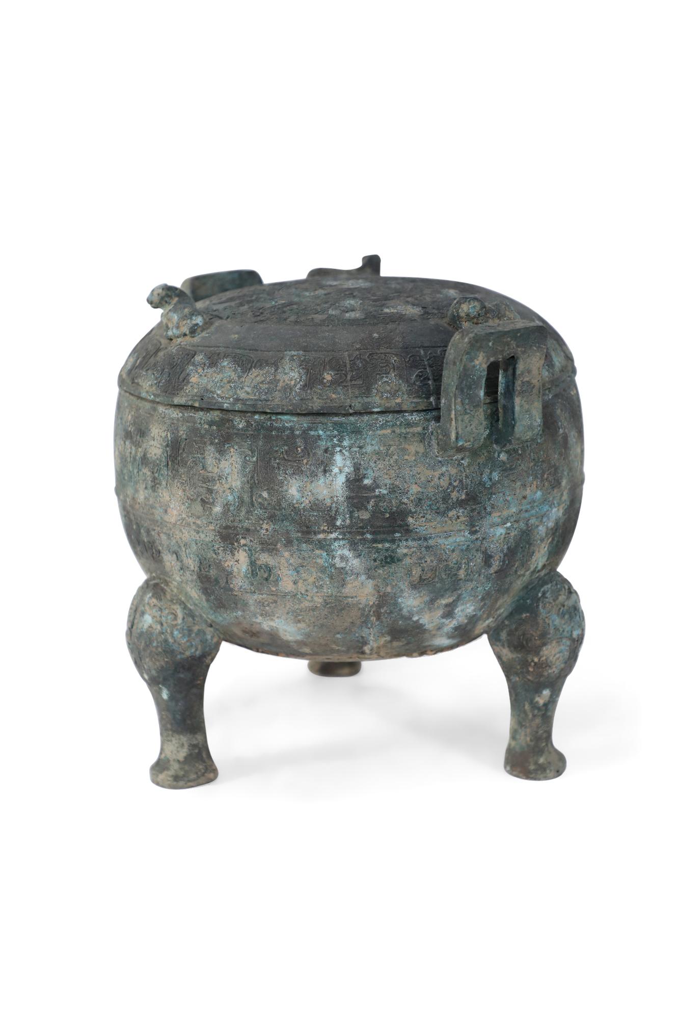 Antique Chinese Han Dynasty-style copy of a patinated bronze pot carved with raised geometric patterned bands wrapping its form, sitting atop three feet, fitted with two handles and topped with a lid bearing three cow figurines.