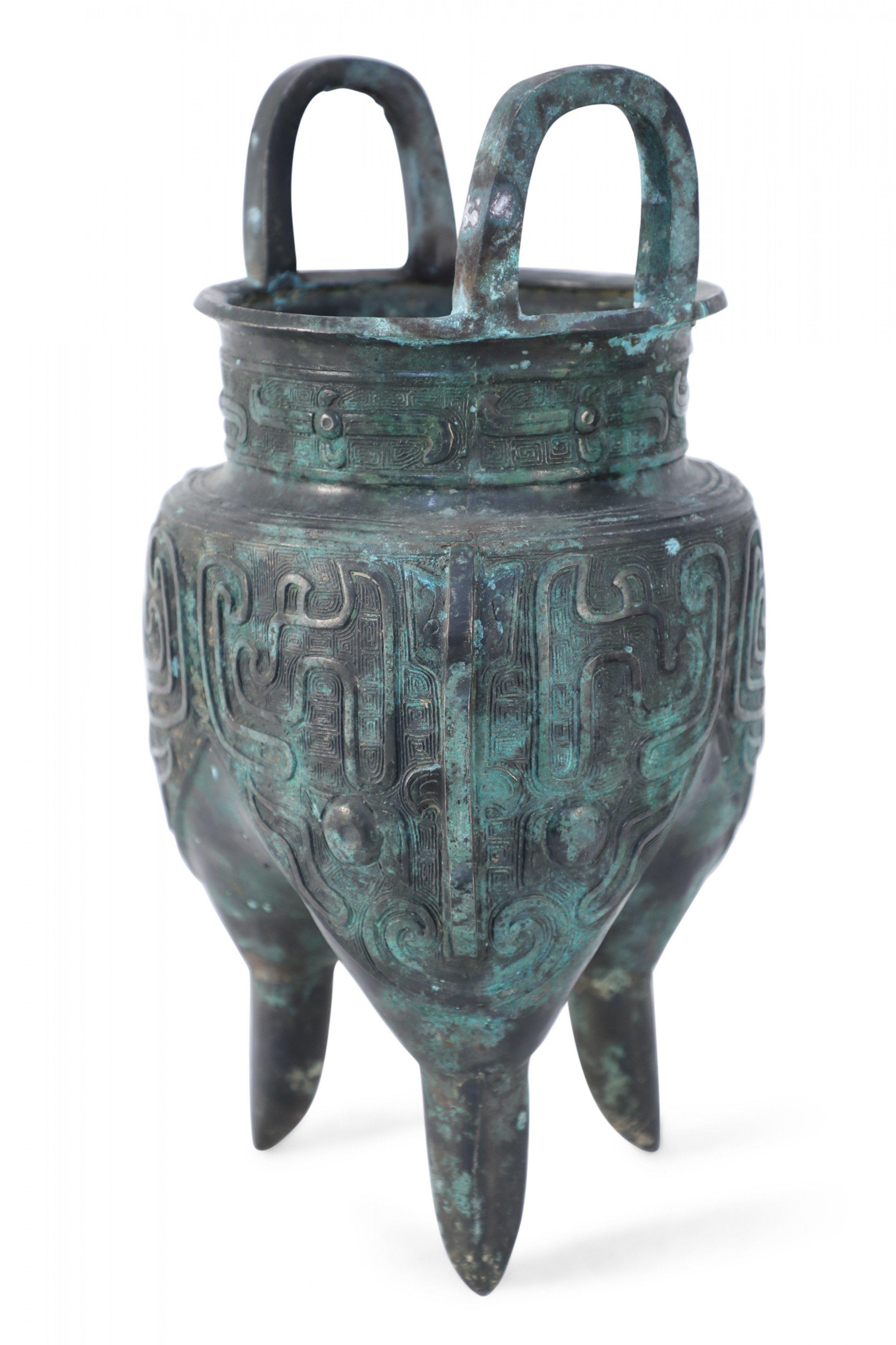 Antica copia in stile Dinastia Han cinese di un recipiente in bronzo patinato intagliato con motivi geometrici e serpentini in rilievo, posto in cima a tre gambe affusolate e rifinito con due manici a forma di C che si estendono verso l'alto.