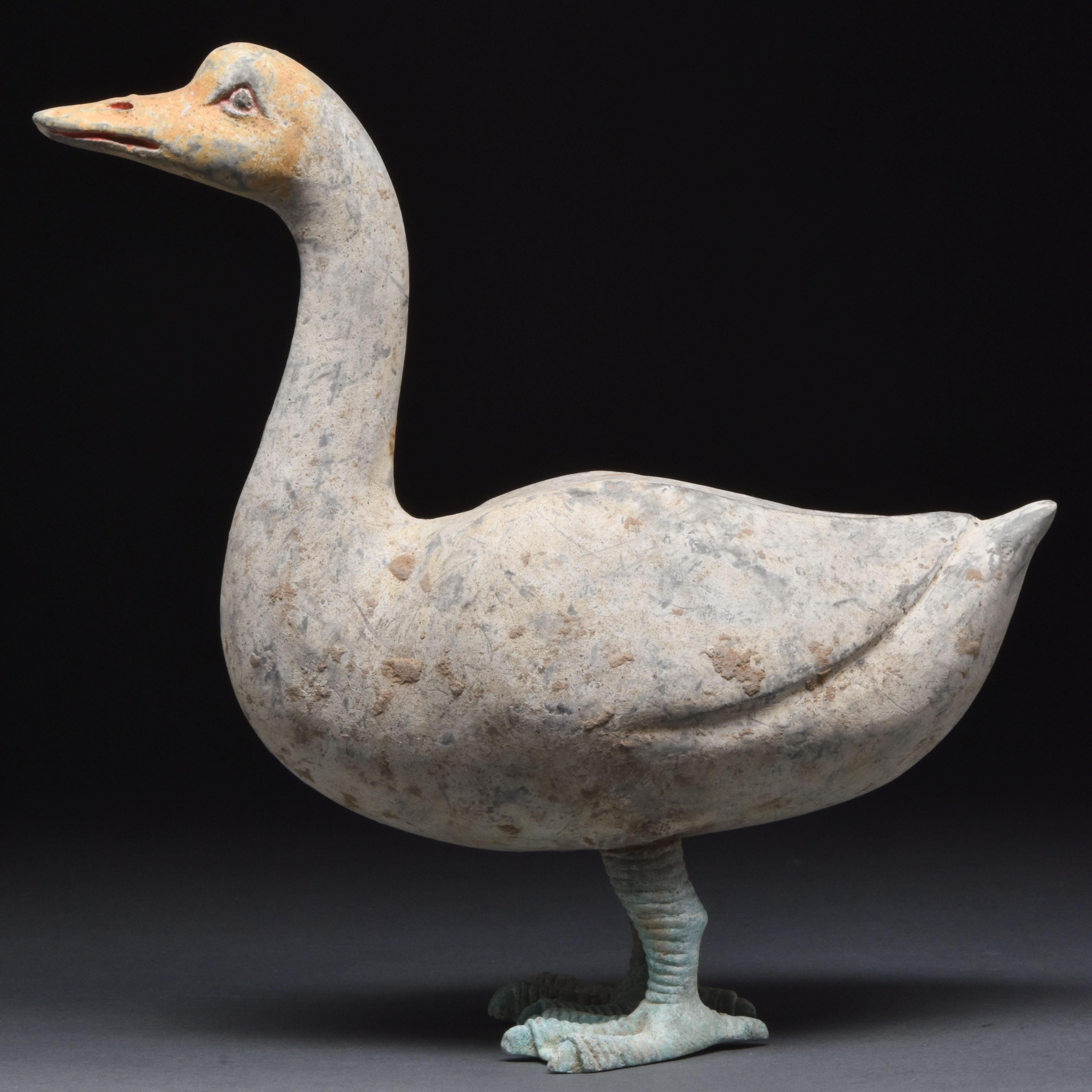 A large ceramic goose standing on its stylised, well-defined bronze legs, which support a white globular body and elegantly slender neck terminating in a yellow-coloured head with a beak and open, attentive eyes. Geese were a significant motif in