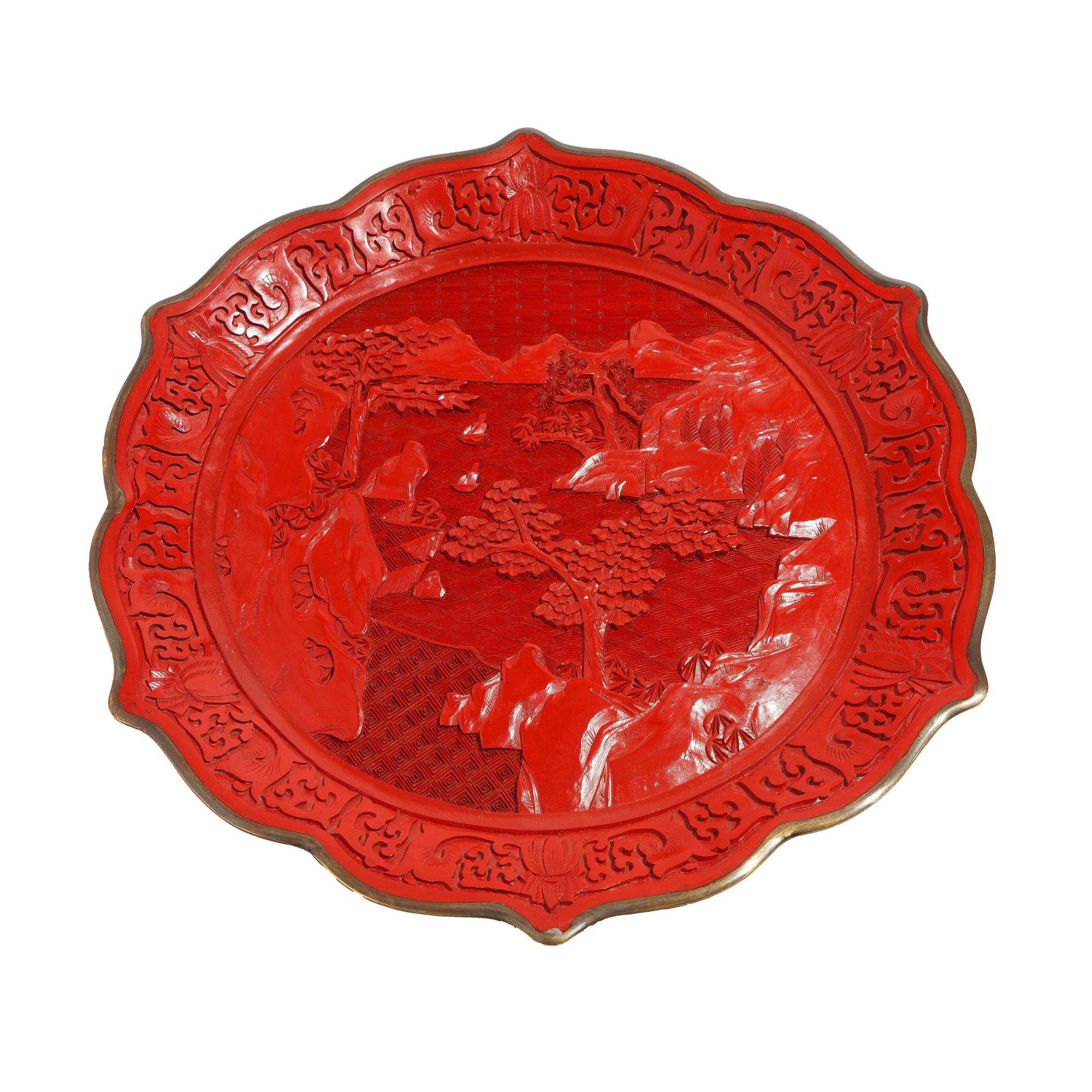 Hand carved cinnabar plate with scalloped rim on brass. The carved scene is of a mountain and river landscape. In the distant background are three undulating mountains facing a body of water with trees growing on promontories.Craggy rock cliffs with
