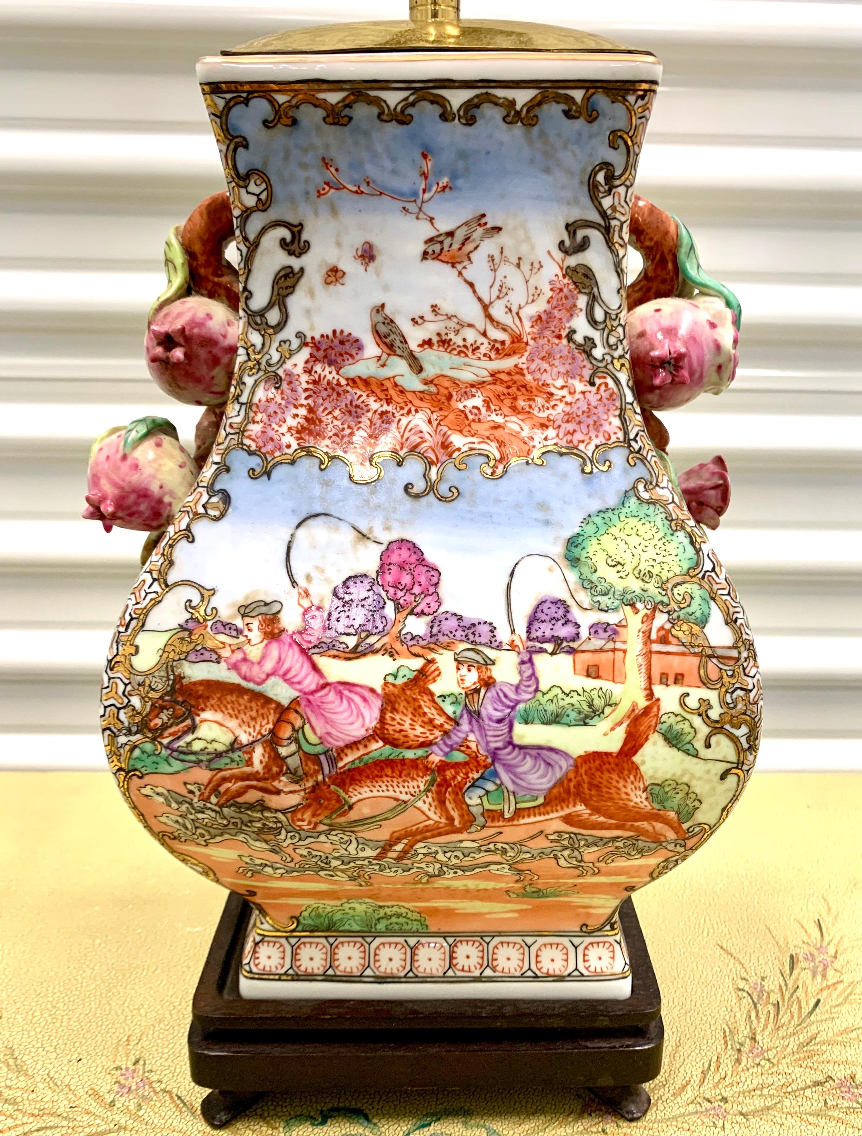 Colorful Chinese export porcelain vase mounted as a lamp with hand painted fox hunt motif. Features raised pomegranate fruit handles with brass hardware and finial. Now electrified and mounted on a wood base. Shade dimensions: 17” W x 12” D x 11.5”