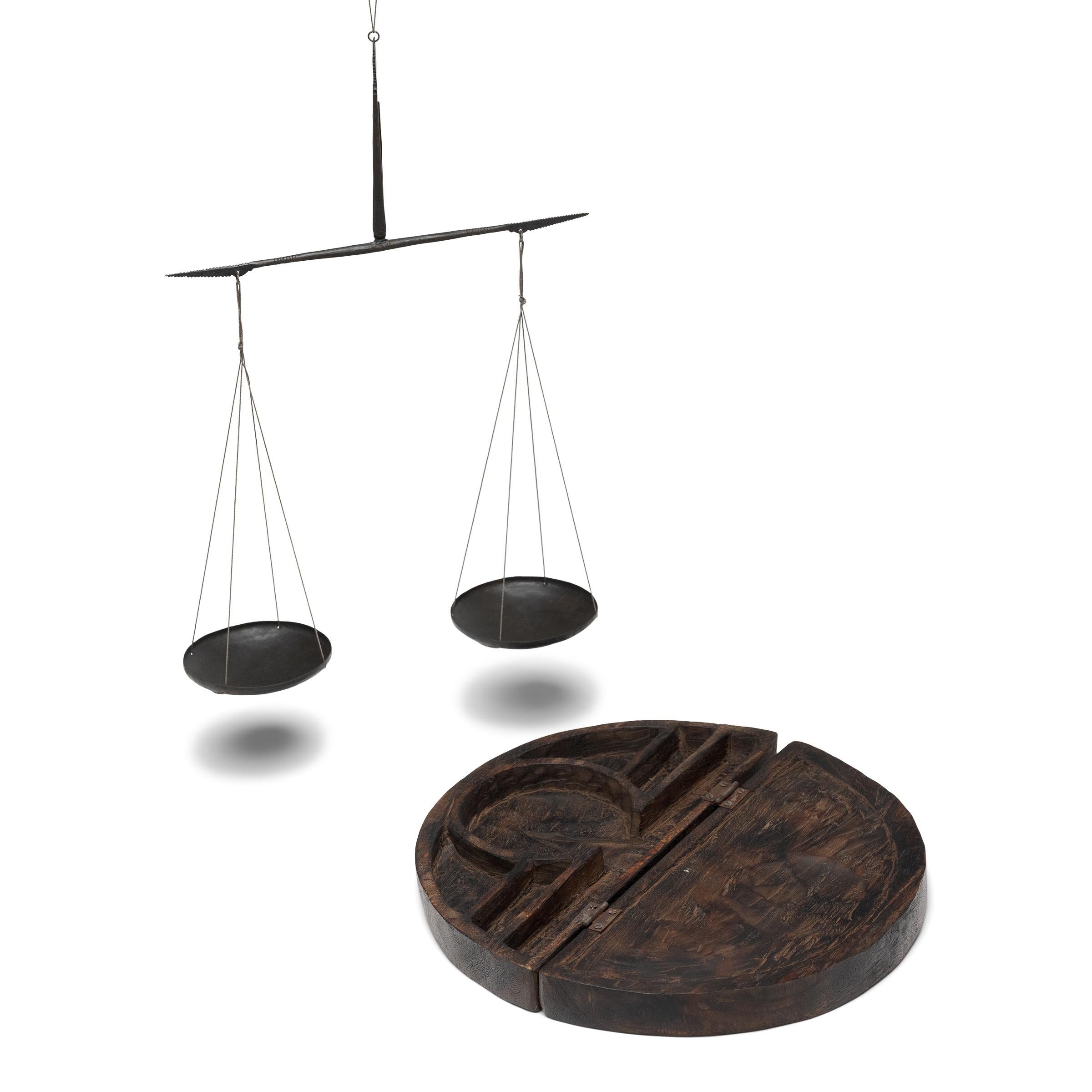 This curious keepsake is a hanging opium scale with brass pans, used to measure opium and other goods by weight. The scale is held in a wooden case that folds to a half-moon shape and is carved in relief with a design of poppy flower bulbs, yet to