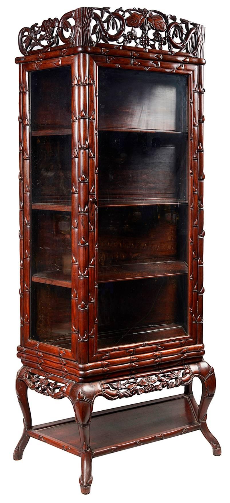 A good quality late 19th century Chinese hardwood display cabinet or vitrine. Having beautifully hand-carved scrolling vine leaves and faux bamboo to the show wood. Having a glazed door opening to reveal two wooden shelves within. Raised on carved