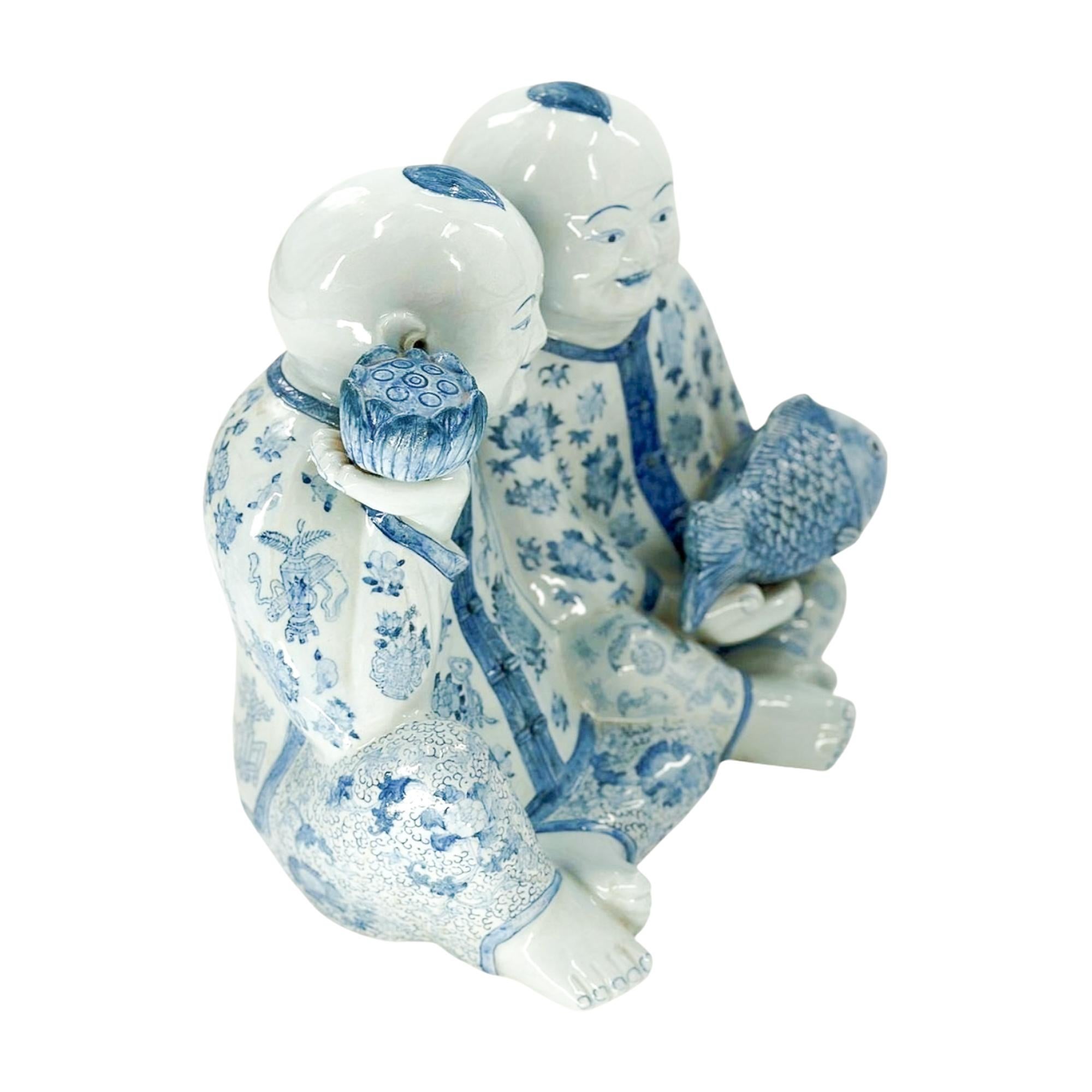 This striking porcelain figure depicts a pair of twin boys rendered in classic blue-and-white, a motif deeply rooted in Chinese decorative tradition. Seated side by side, one child holds a lotus blossom while the other carries a large fish, both