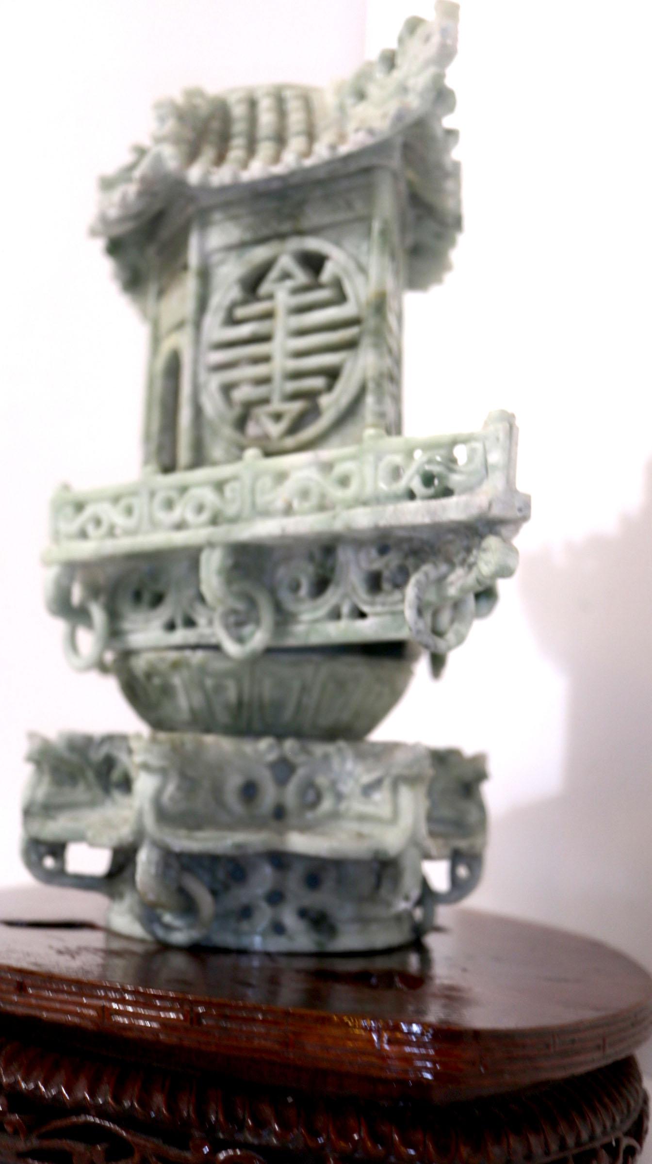 This is a special honan jade temple carving censer with a small Buddha figure visible from inside the carving in white. The color is cool with hints of white. It rests on an oval rosewood-colored base that is also carved around the edges.
In