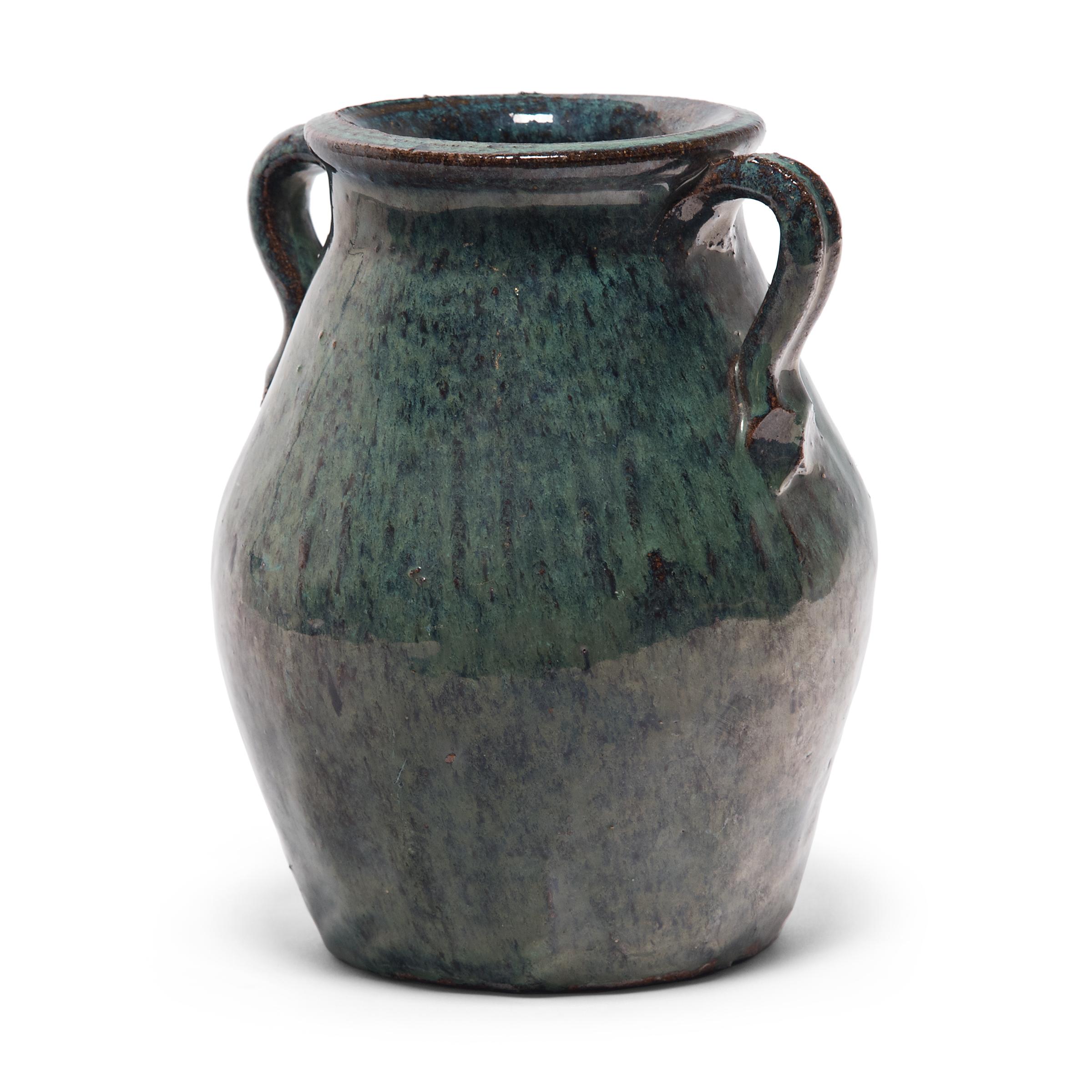 A luminous blue-green glaze sheets, pools and drips down the surface of this early 20th century kitchen jar. The pear-shaped jar has a tapered body and a wide flared mouth framed by simple strap handles. The monochrome jade green glaze exhibits a
