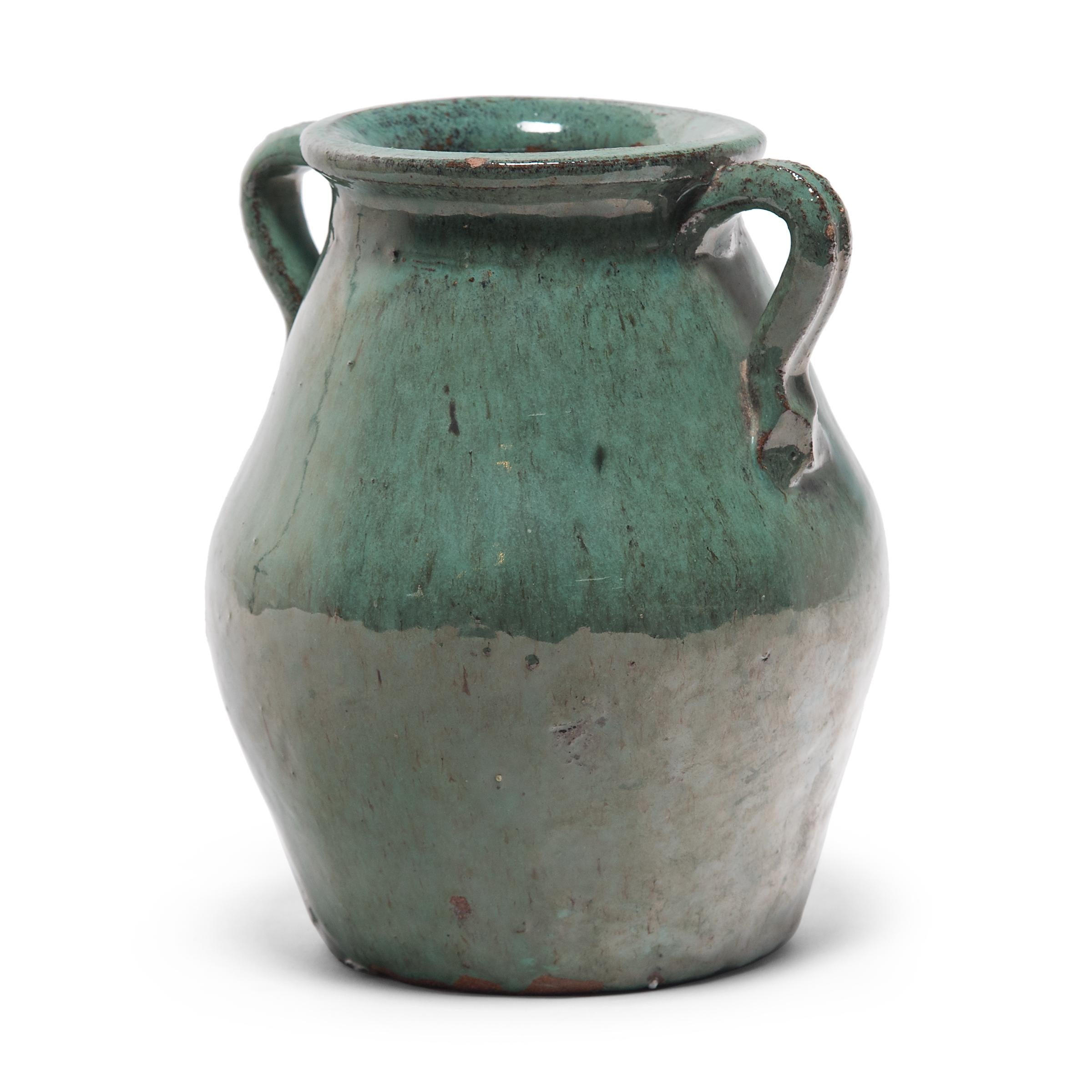 A luminous blue-green glaze sheets, pools and drips down the surface of this early 20th century kitchen jar. The pear-shaped jar has a tapered body and a wide flared mouth framed by simple strap handles. The monochrome jade green glaze exhibits a