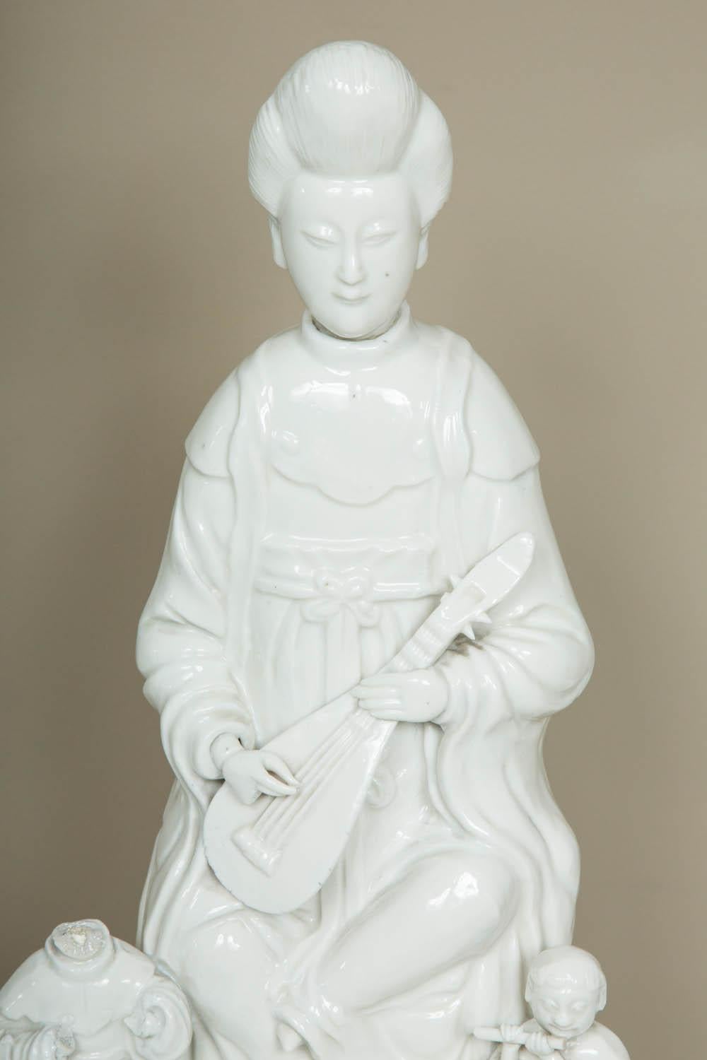 A Kangxi period (1662-1722) Blanc de Chine porcelain model of a seated Guanyin playing a music instrument, at her side two children playing the flute and wearing european costumes; from the kilns in Dehua, Fujian Province.
Condition: neck reglued