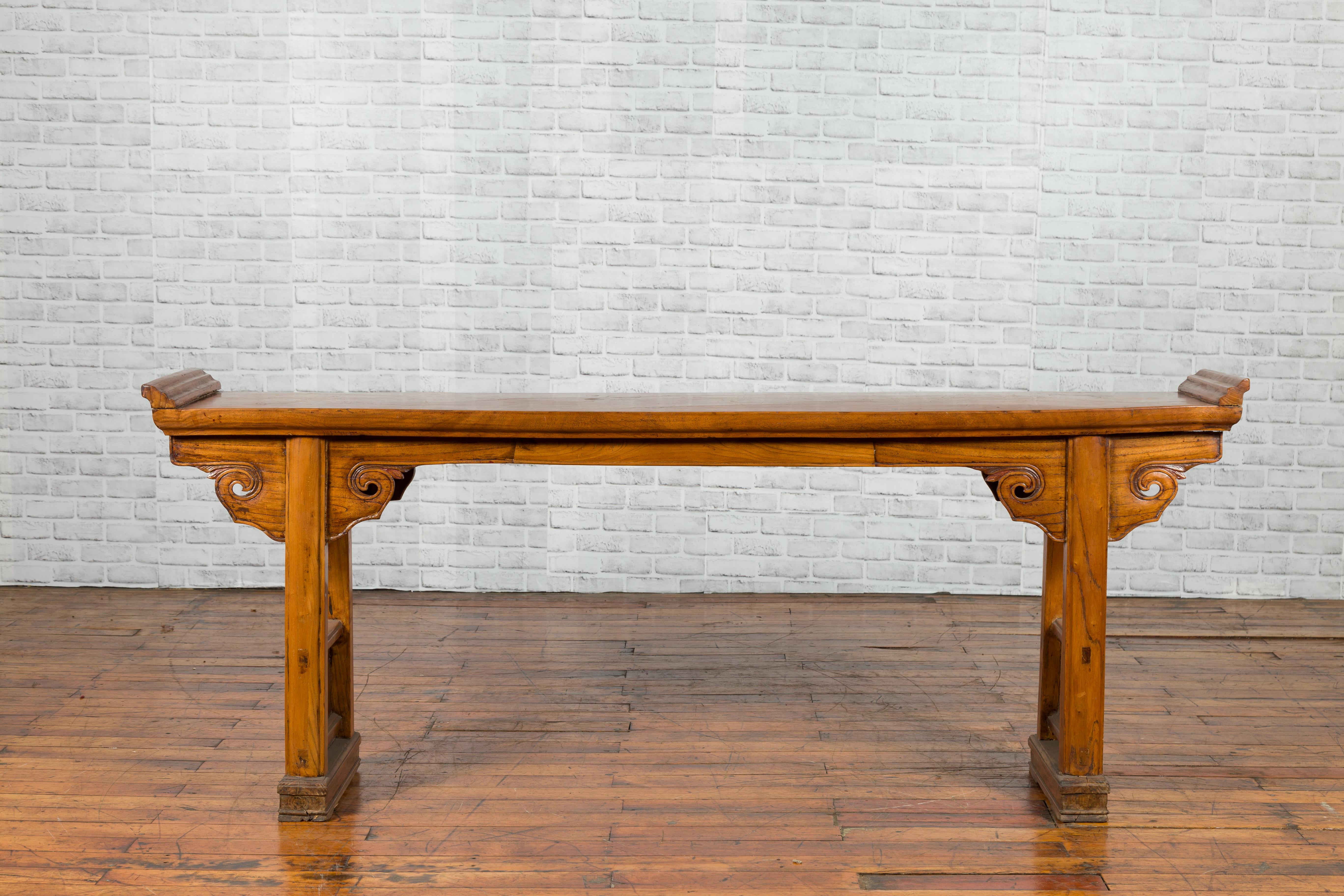 A Chinese Qing dynasty period elm altar console table from the late 19th century, with everted flanges, carved cloud spandrels and pierced motifs. Created in China during the later years of the 19th century, this elm console table features a long