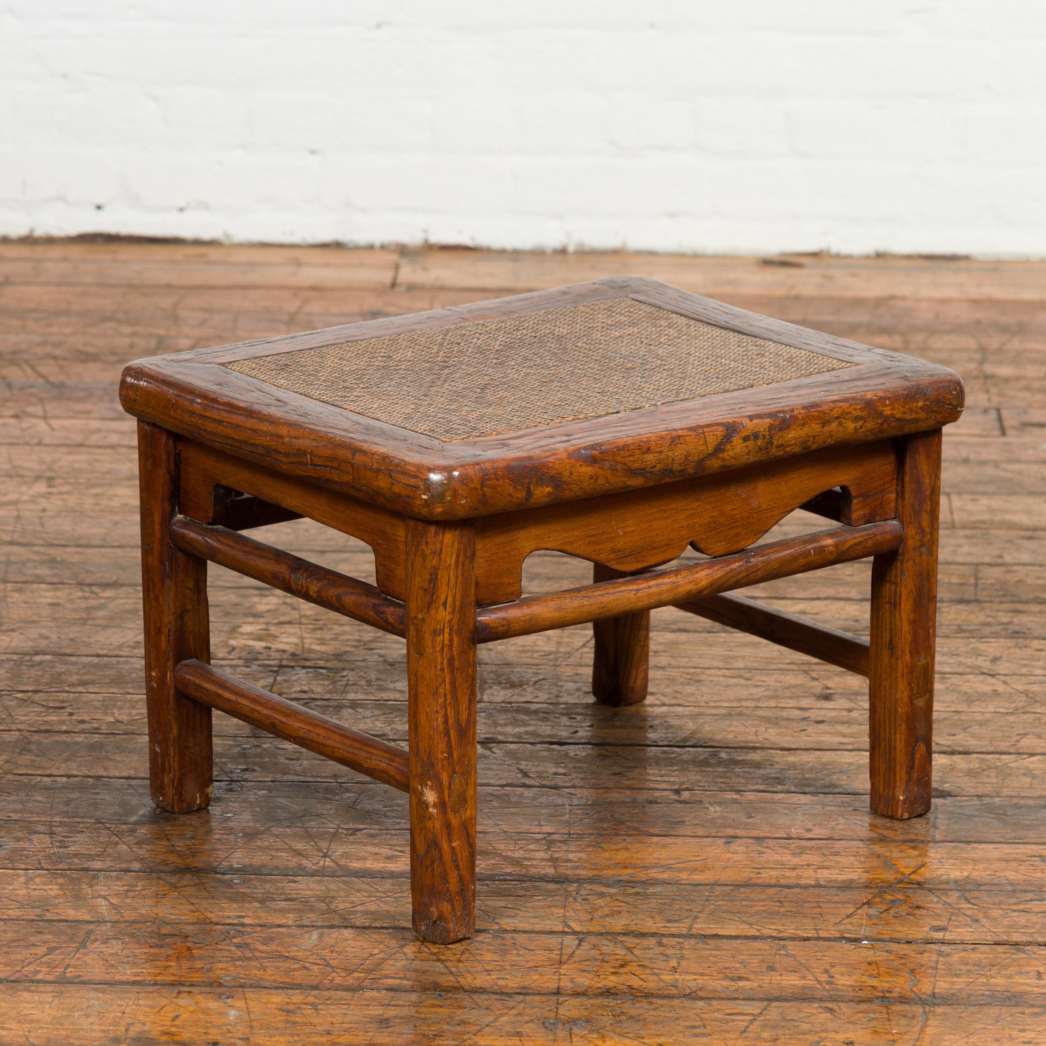 A Chinese late Qing Dynasty period footstool from the early 20th century with rattan top, carved apron and side stretchers. Discover the perfect blend of function and art in this early 20th-century Chinese footstool from the late Qing Dynasty