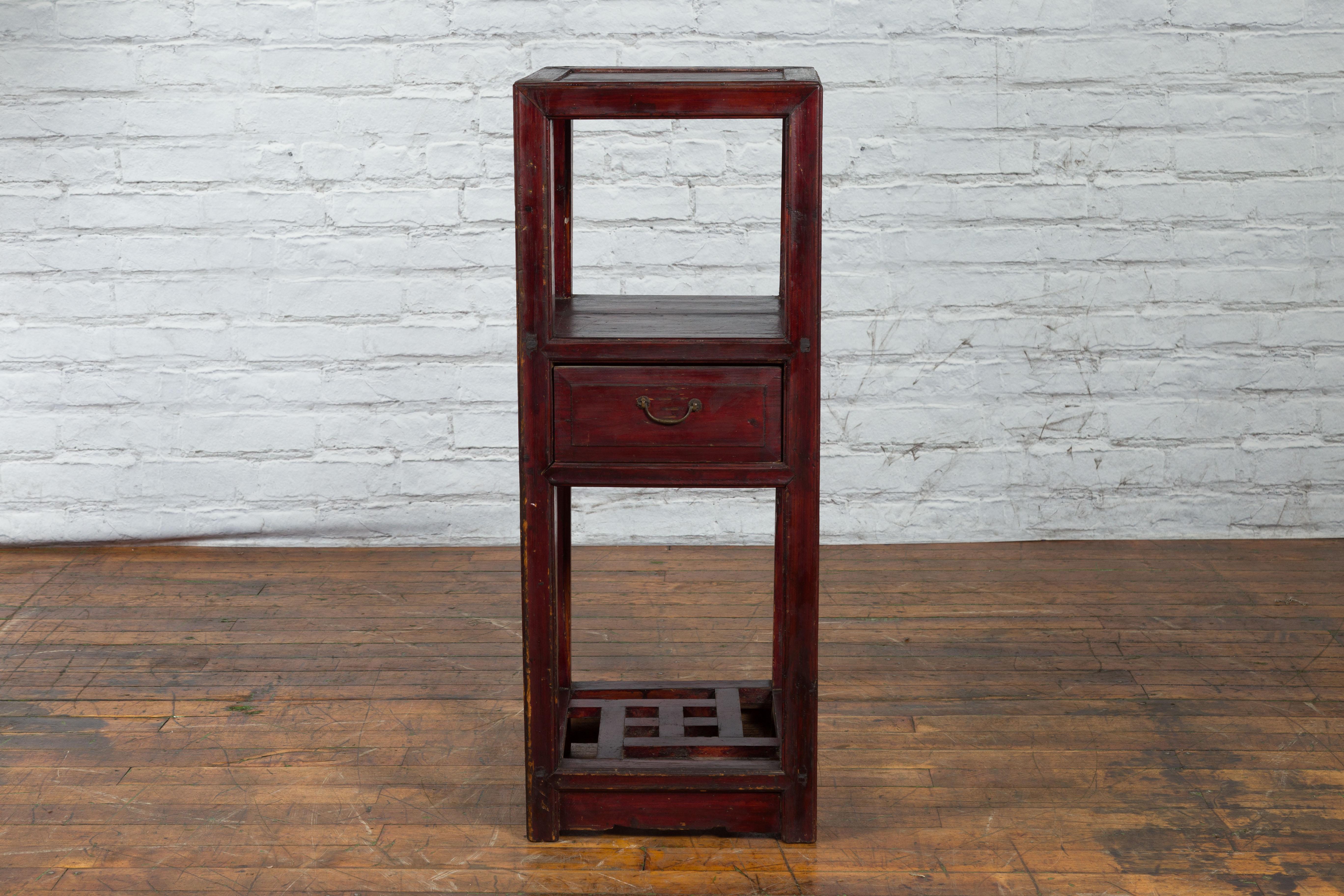 A Chinese late Qing Dynasty period tiered side table from the early 20th century with reddish brown lacquer, carved fretwork shelf and single drawer. Created in China during the late Qing Dynasty period in the early years of the 20th century, this