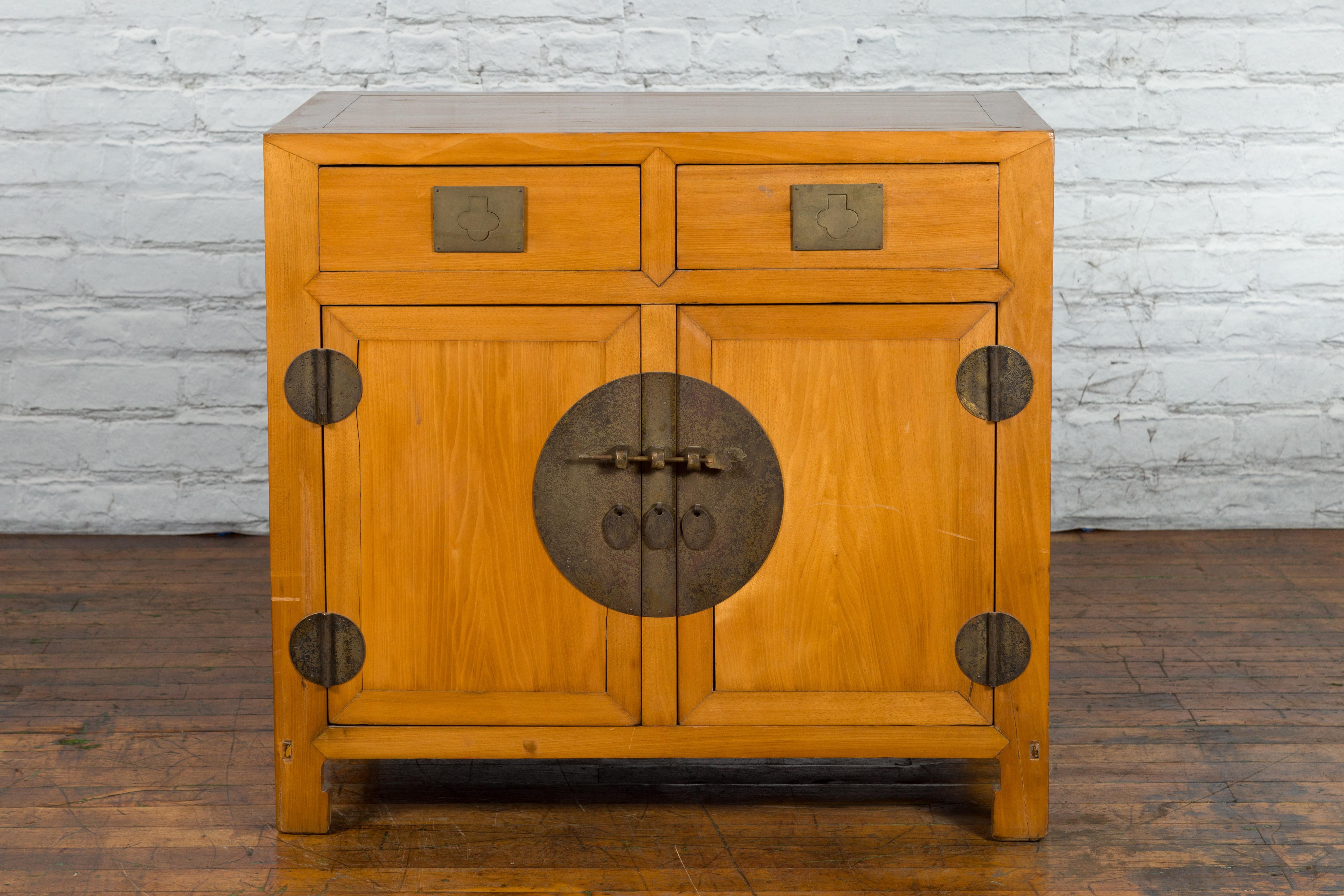 A Chinese Late Qing Dynasty period elm wood cabinet from the early 20th century, with brass medallion, teardrop pulls, two drawers and two doors on horse hoof feet. Created in China during the later years of the Qing Dynasty in the early 20th