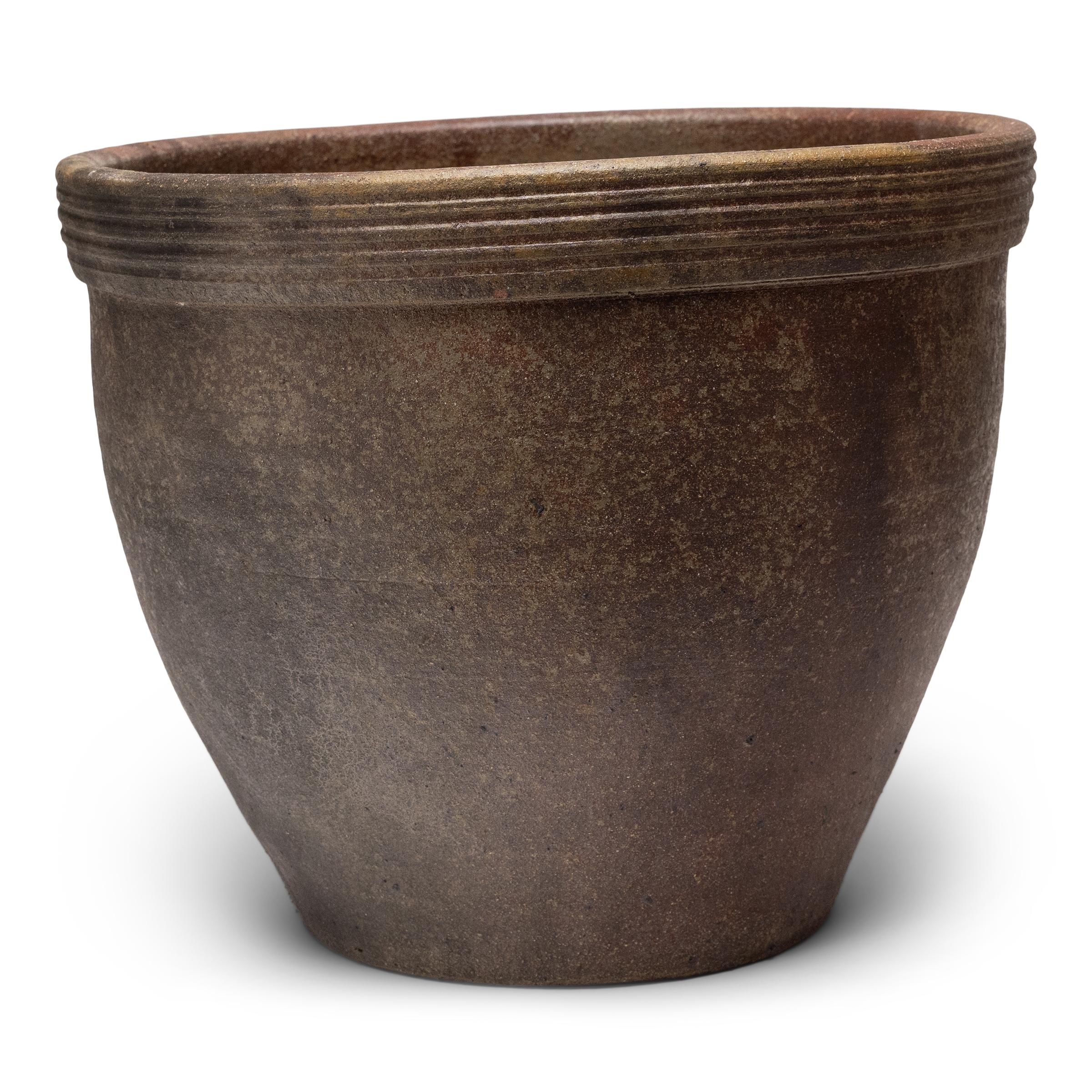 Questa piccola fioriera in terracotta ha una forma minimale e una finitura splendidamente irregolare. Il vaso in ceramica ha una silhouette affusolata, con una bocca larga, una base stretta e un ampio bordo con creste orizzontali. Lasciato senza
