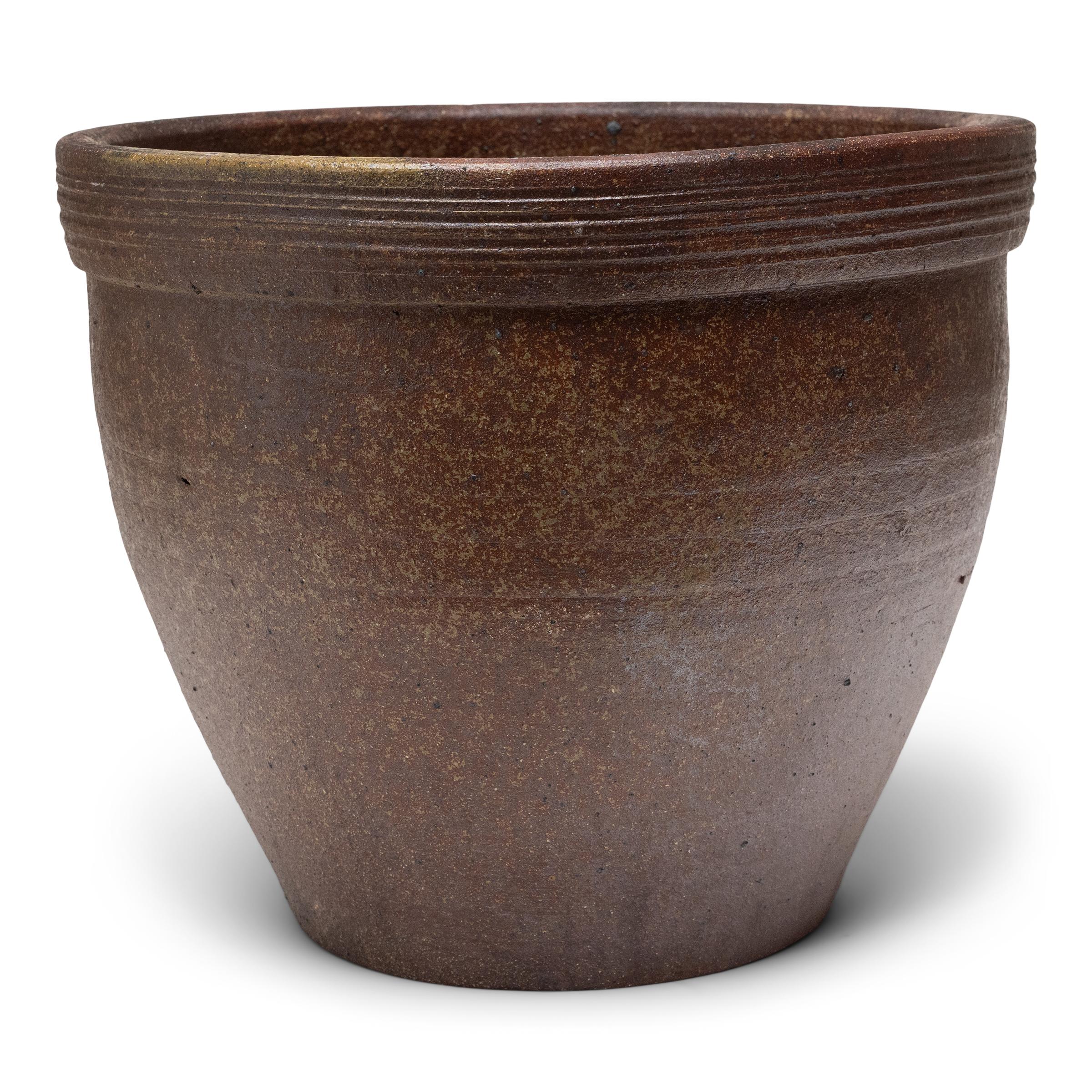 Esta pequeña jardinera de terracota tiene una forma minimalista y un bonito acabado irregular. El recipiente de cerámica tiene una silueta ahusada, con boca ancha, base estrecha y borde ancho con estrías horizontales. Si se deja sin decorar, el