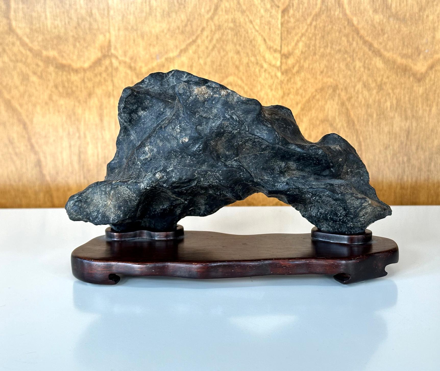 A small desk top Chinese scholar stones (also known as Gong Shi, meditation stone and spirit rock) fitted on a hand-carved wood stand circa late 19th century. The specimen is of a black Lingbi type stone in an arch form mimicking a horizontal