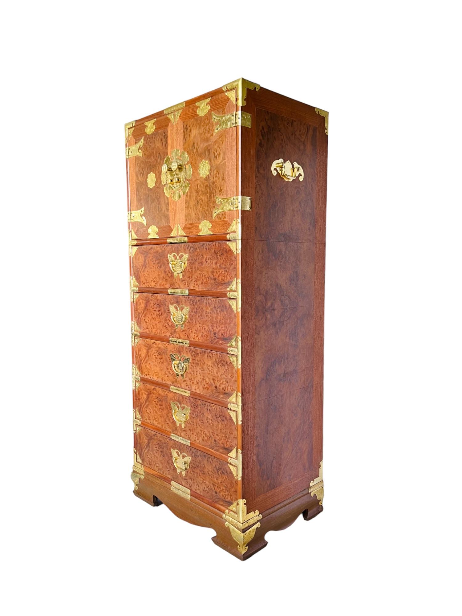 A vintage 1960s Chinese Campaign style lingerie chest finely crafted of teak and burl elm wood and embellished with etched brass mounts. This stunning chiffonier features a medallion adorned upper cabinet and boasts a total of eleven drawers - six