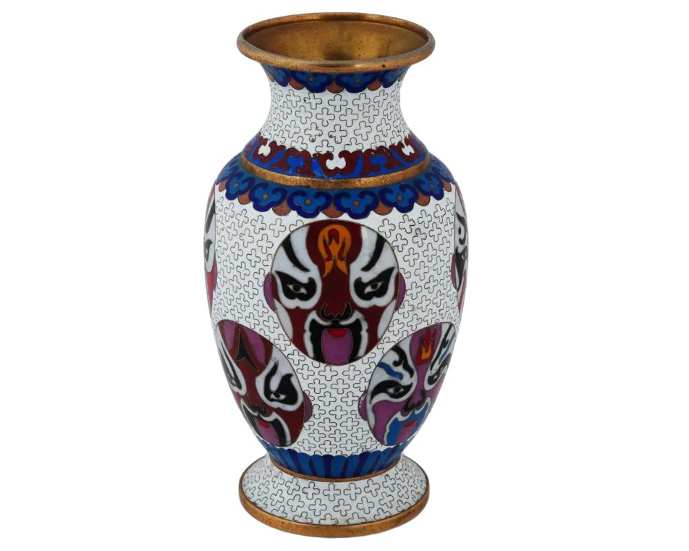 A vintage Chinese footed enamel over brass vase. The urn shaped vase is adorned with a polychrome enamel design of masks surrounded by a traditional ornament on the white ground made in the Cloisonne technique. The neck features foliage and