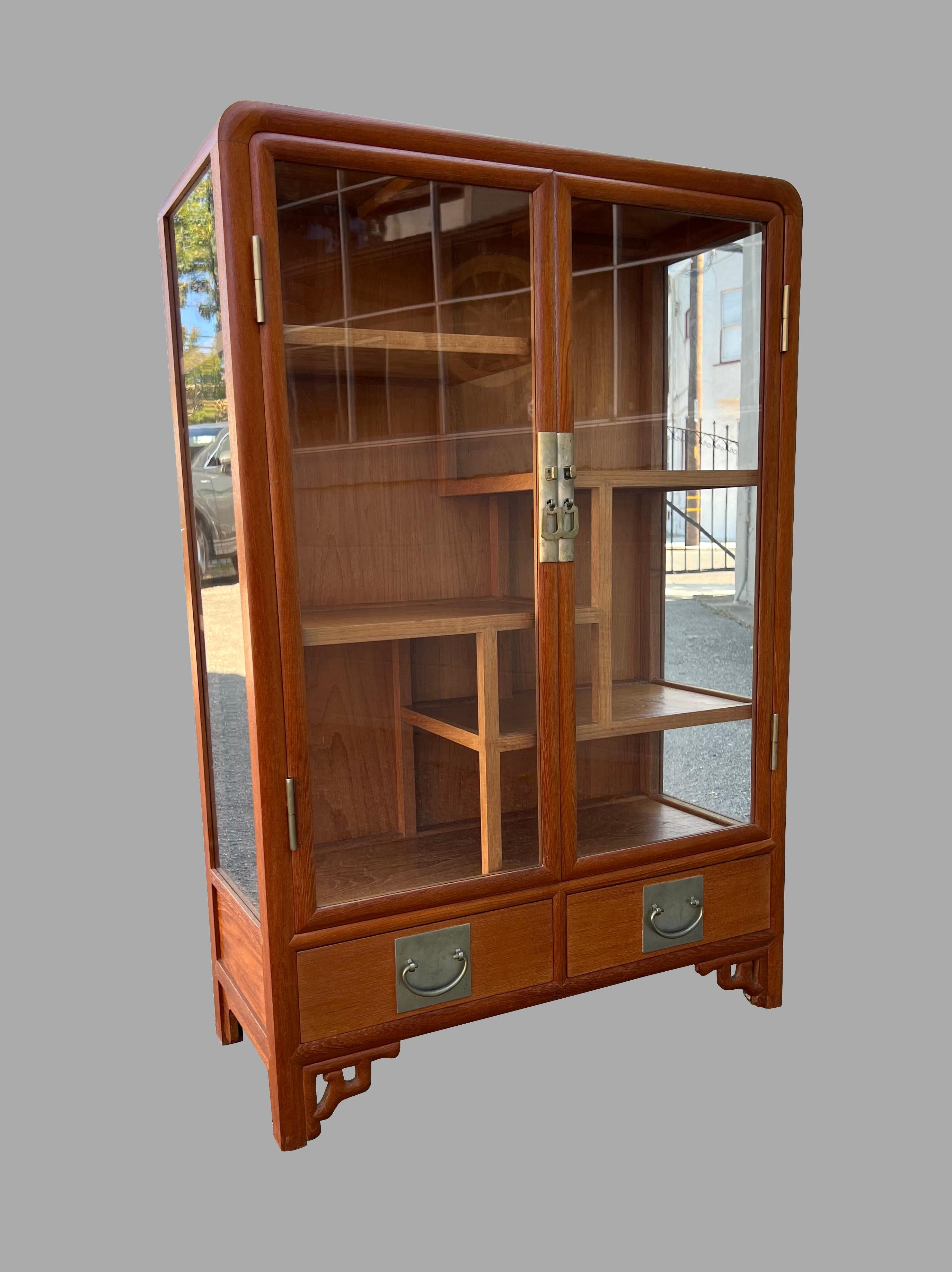 A small scale Chinese teak display cabinet of compact proportions, the glazed double doors over 2 short  dovetailed drawers resting on Asian style open bracket feet. Retains its original white brass hardware. Lock not present. Circa 1930-1950.