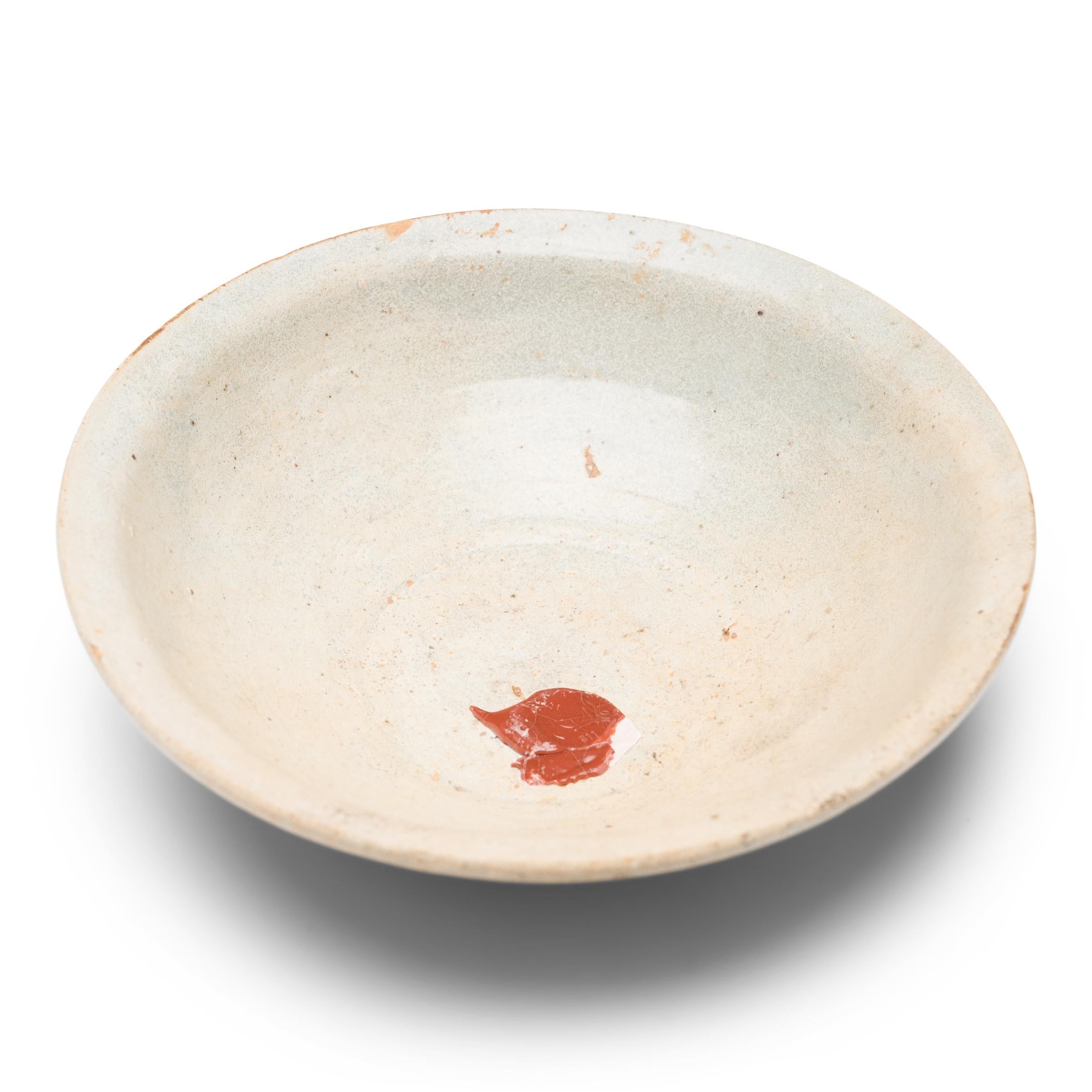 This lovely earthenware bowl from the early Ming dynasty (1368-1644) is cloaked in an allover celadon glaze with smooth texture and an opaque, clouded finish. Once used daily for food and drink, the shallow bowl is potted with a short, footed base