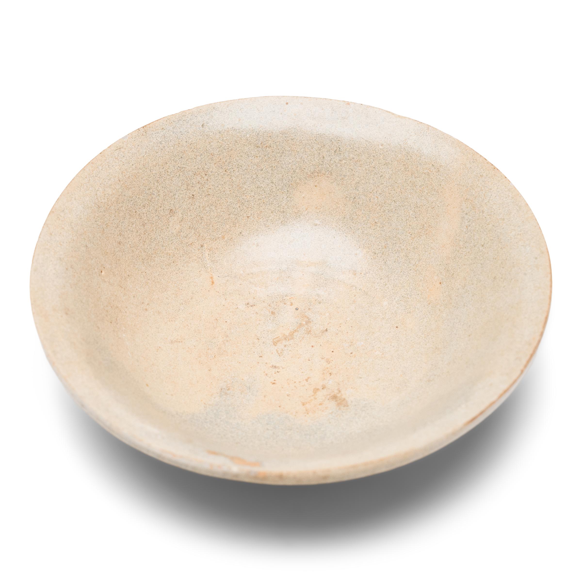 This lovely earthenware bowl from the early Ming dynasty (1368-1644) is cloaked in an allover celadon glaze with smooth texture and an opaque, clouded finish. Once used daily for food and drink, the shallow bowl is potted with a short, footed base