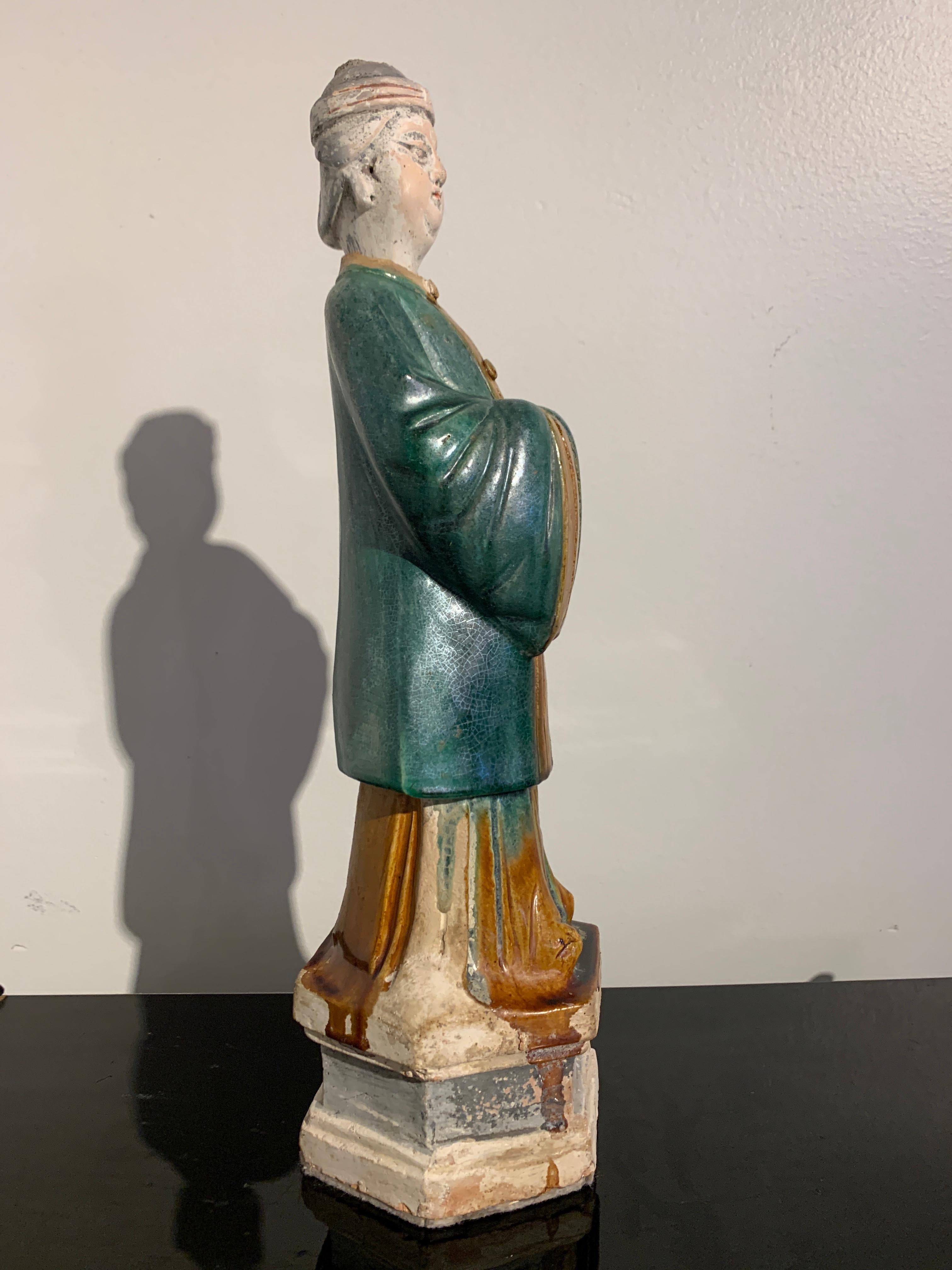 A tall Chinese green and amber glazed figure of an attendant, Ming Dynasty, 16th-17th century, China. 

The figure stands upon an integral plinth, dressed in long robes and a generous overcoat with wide sleeves, hands hidden within the folded