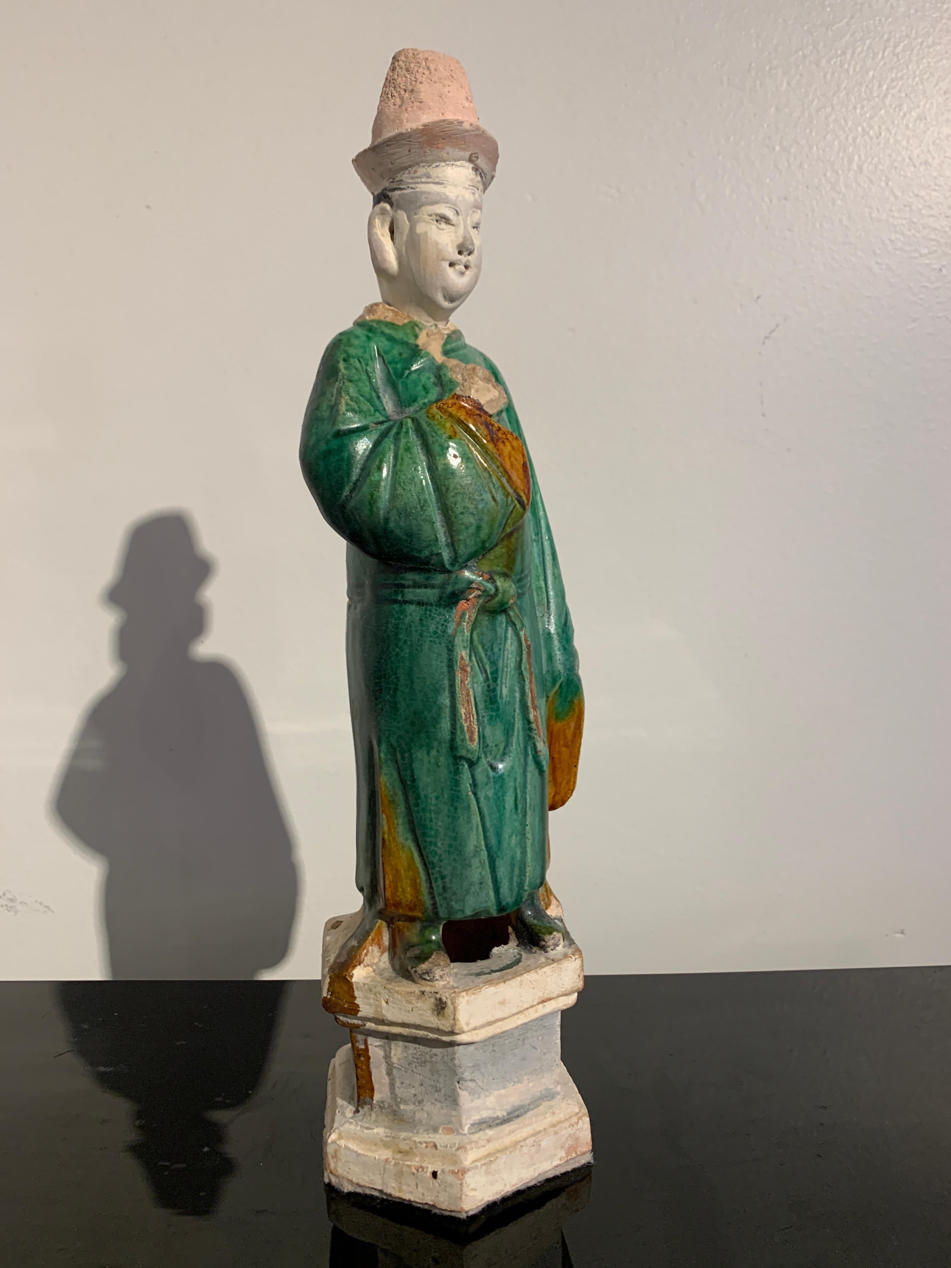 A nicely sized Chinese green and amber glazed pottery figure of a male attendant, Ming Dynasty, 16th-17th century, China.

The figure stands upon a tall integral hexagonal plinth, and is dressed in oversized green glazed robes with amber glazed