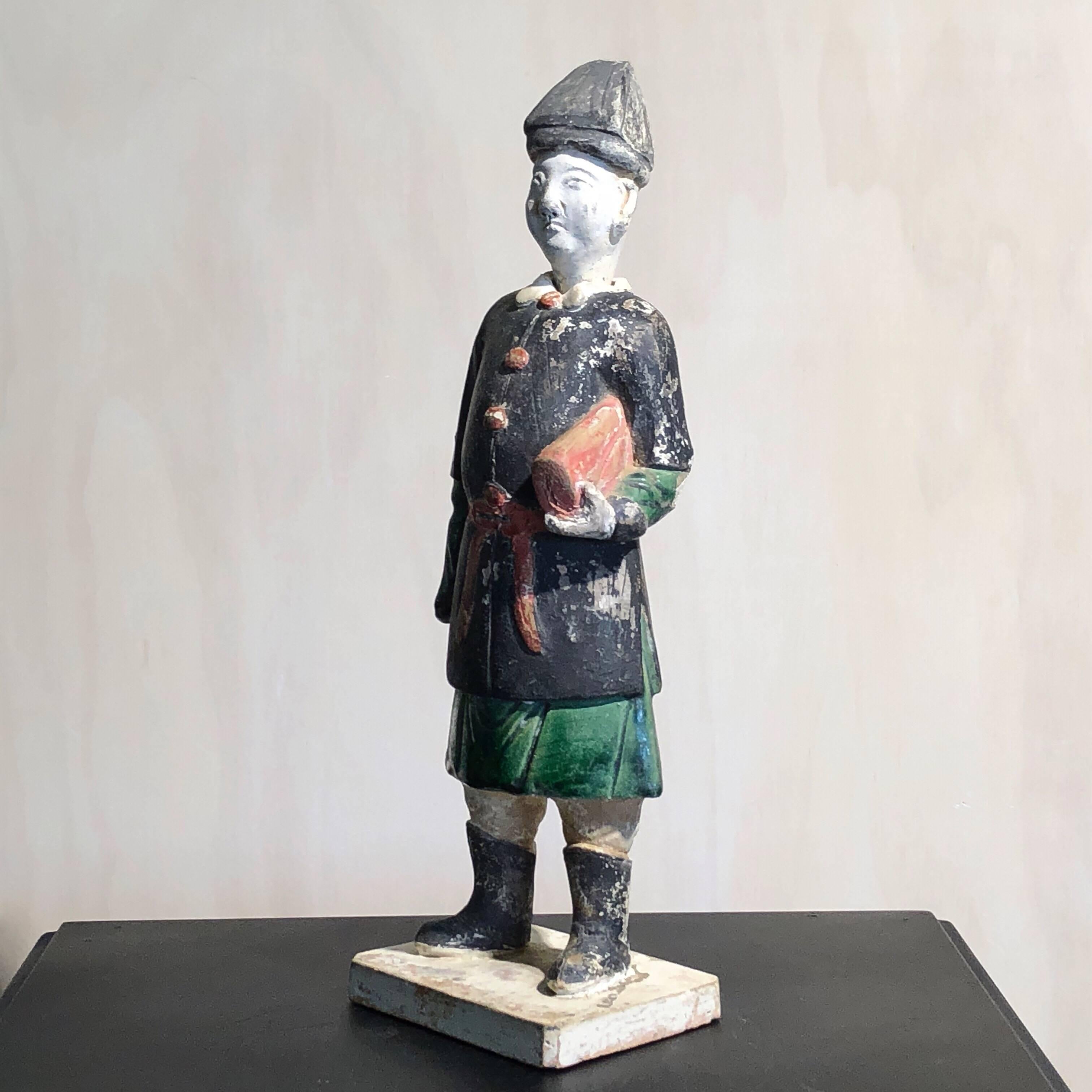 Chinese pottery official, the partially glazed figure standing with bundle of goods beneath one arm, wearing a black painted overcoat with red buttons and sash over a green glazed tunic, his head separate.

