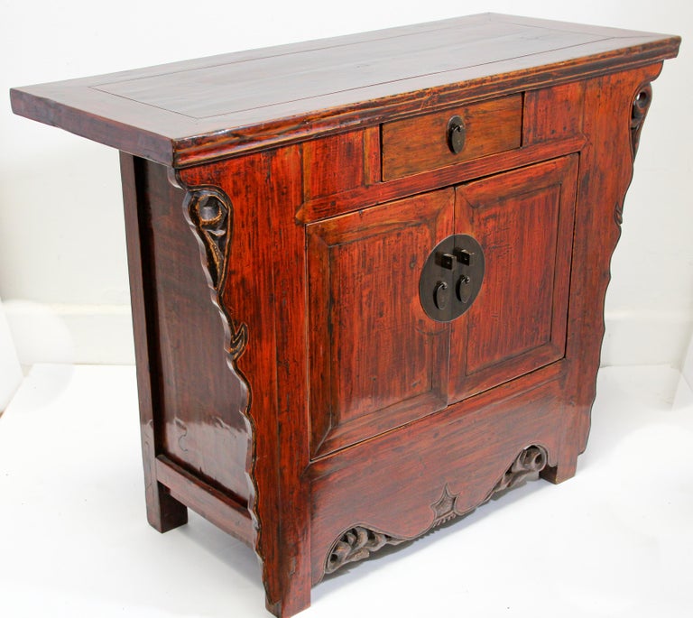 Chinese Ming style hand painted red and brown lacquered cabinet with carved sides, drawer and doors.
The piece features intricate hand carved details and an antique look patina. 
Behind its two doors is ample storage, including a single