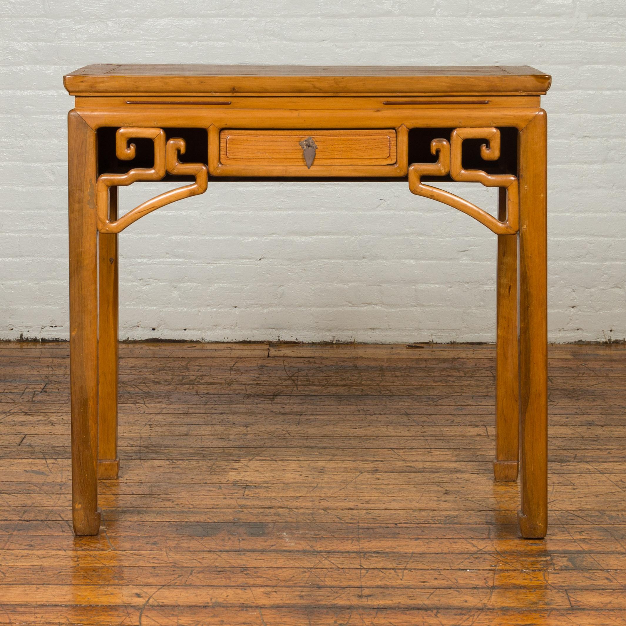 A Chinese Ming Dynasty style desk table with carved apron, single drawer and horse hoofed legs. Born in China, this Ming style table features a rectangular waisted top sitting above a single drawer flanked with carved geometric spandrels. Raised on
