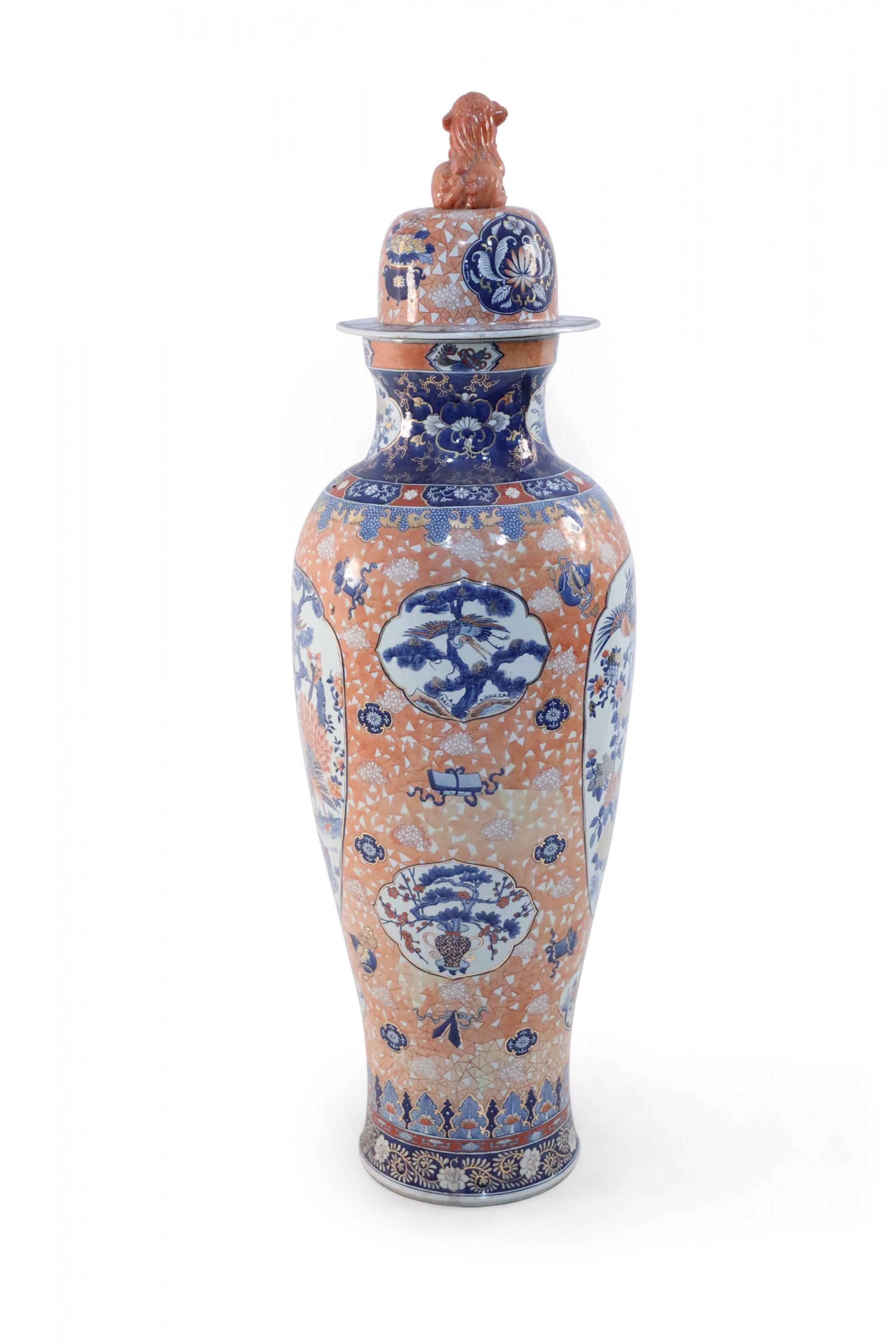 Chinese monumental, lidded porcelain urn celebrating Japanese Imari ware in both palette and design, featuring natural scenes of birds and flowering trees set against a light-orange, geometric-patterned background, and topped with a foo dog-finial