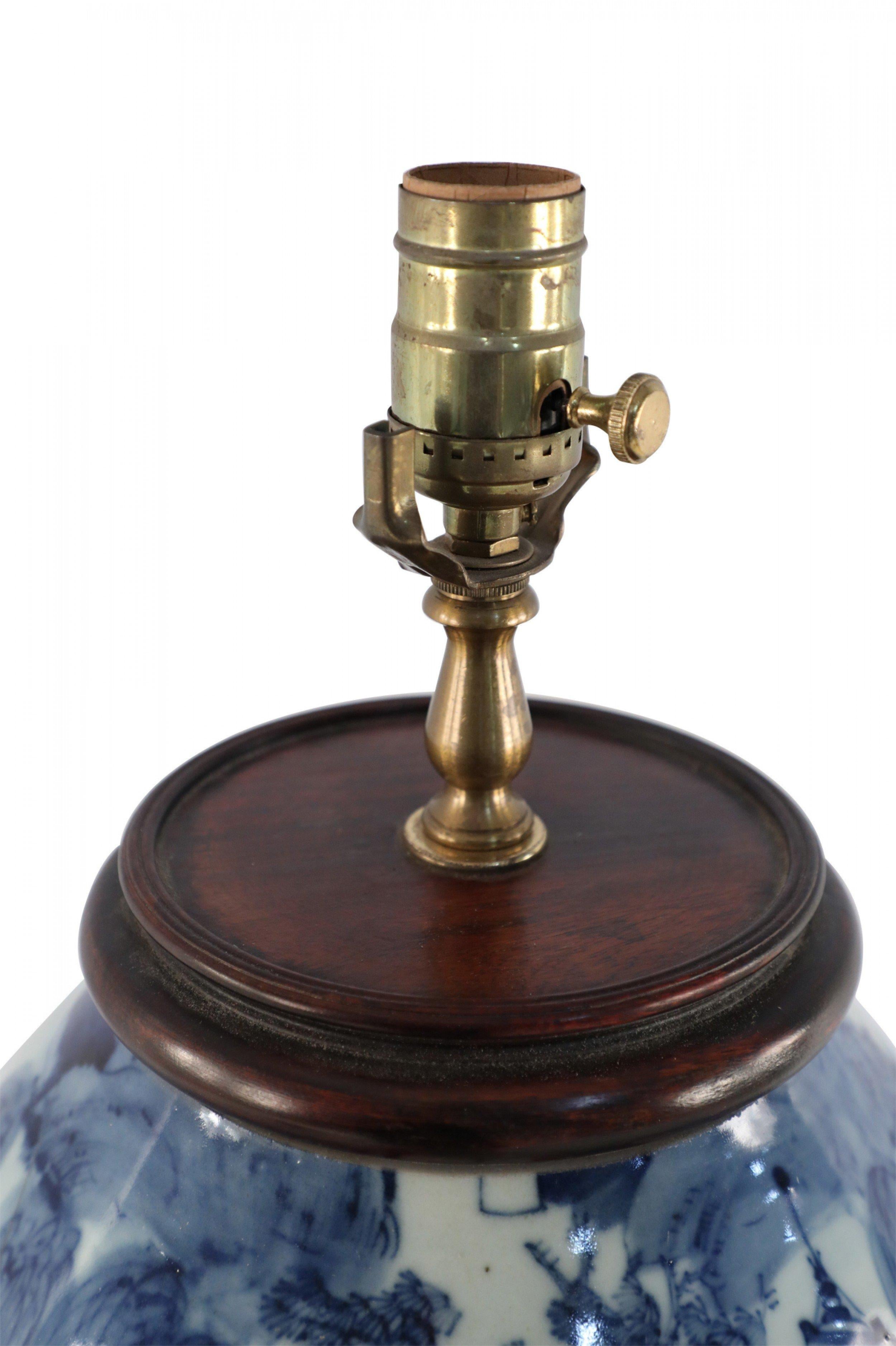 Antique Chinese (Early 20th Century) table lamp made from an off-white and blue, baluster-shaped, porcelain vase capturing a vibrant village along a river, mounted on a brown wooden base with brass hardware.
