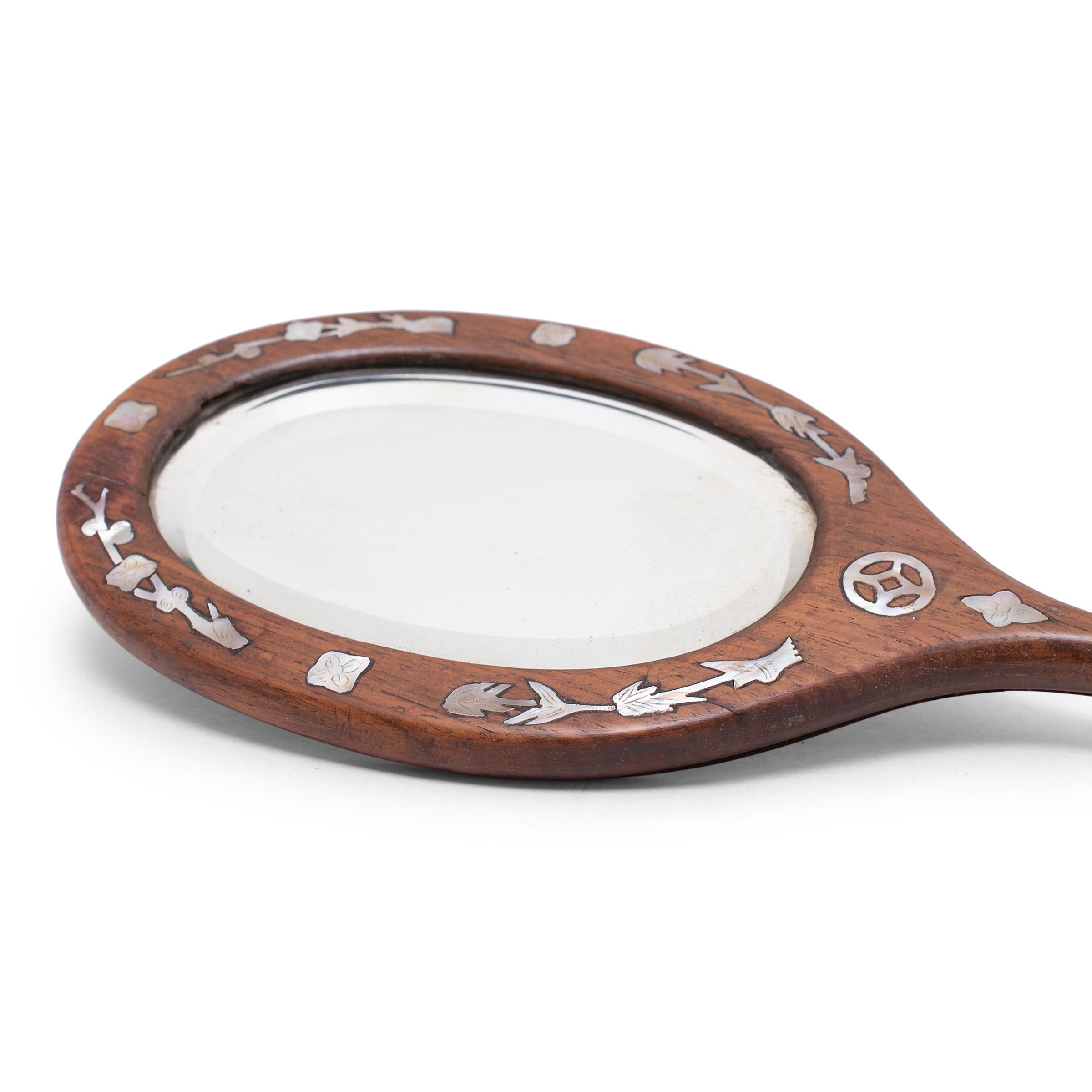 Dieser ovale Handspiegel aus dem frühen 20. Jahrhundert ist in einen edlen Hartholzrahmen gefasst, der mit Perlmuttintarsien aus Bambushalmen und Pflaumenblütenzweigen verziert ist. Der Griff ist mit einer Münze für Reichtum und einem Shou-Symbol