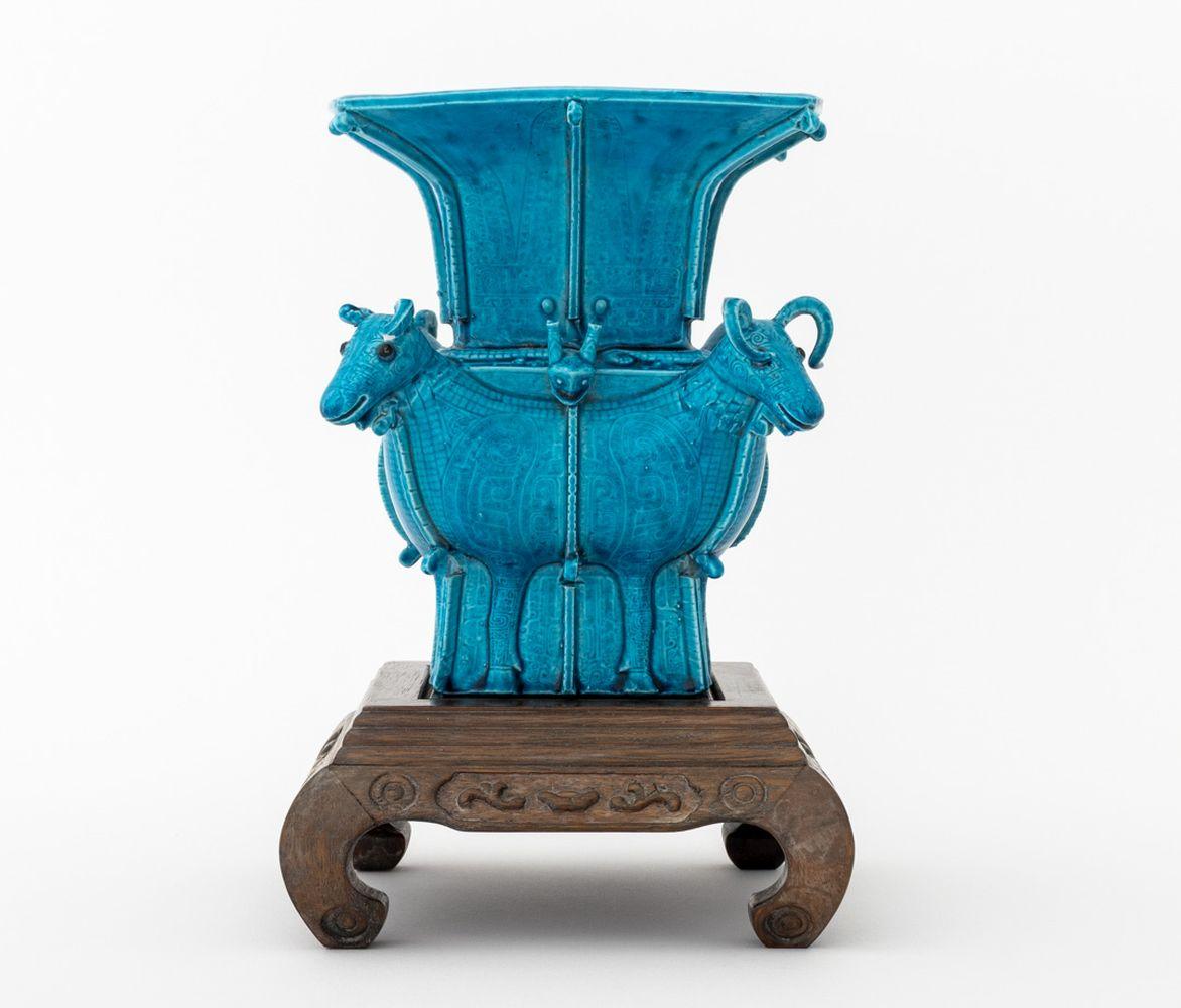 Chinesische Vase mit Widderkopfmotiv aus pfauenblau glasiertem Porzellan, archaisch, mit Sechs-Zeichen-Siegelmarke auf der Unterseite, zusammen mit geschnitztem Holzständer. Vase: 7,75