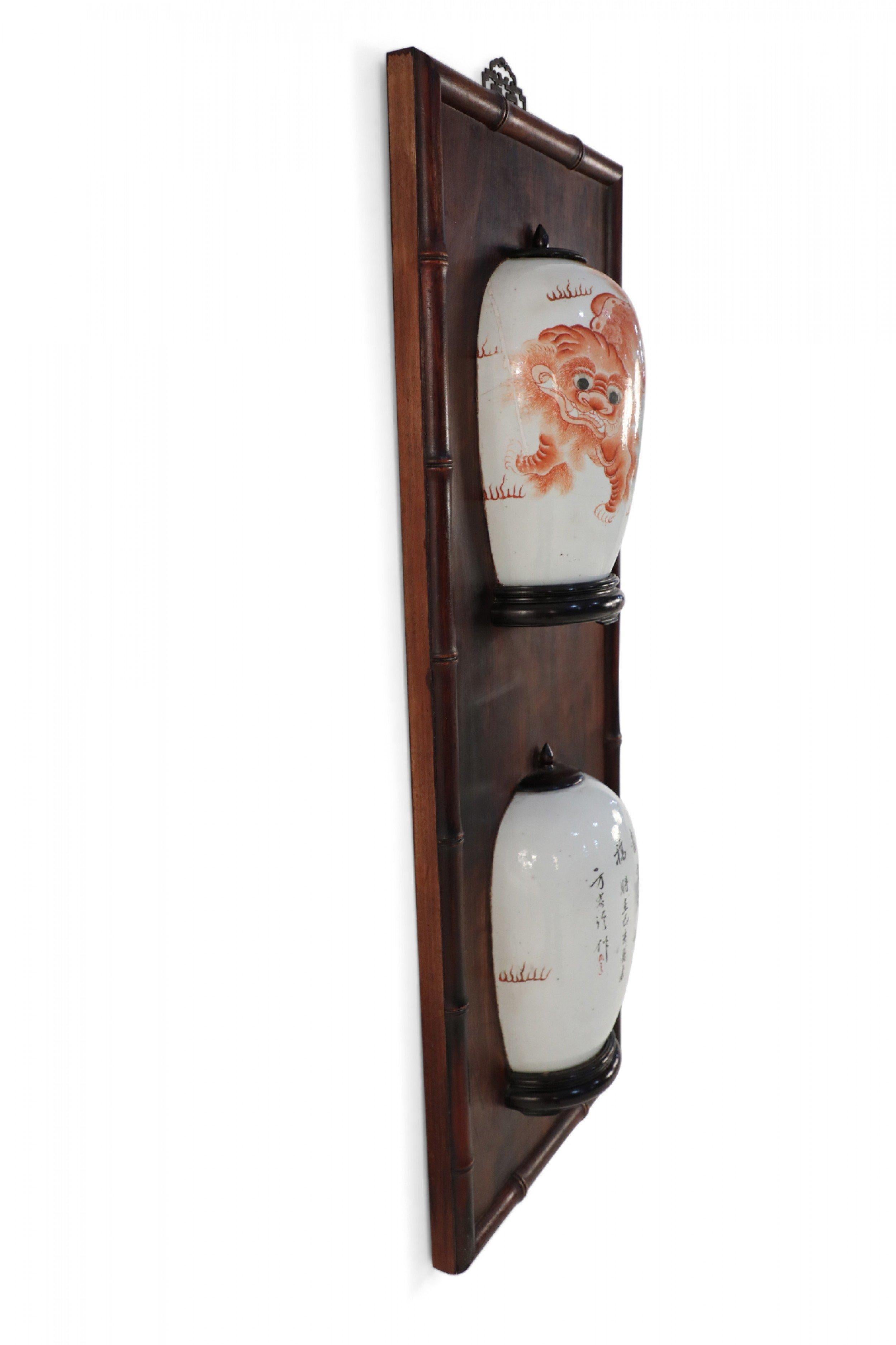 Antigua placa de pared china (finales del siglo XIX) que enmarca una jarra bisecada con tapa de porcelana pintada con perros foo de color naranja y personajes en madera con un borde decorativo de imitación de bambú.