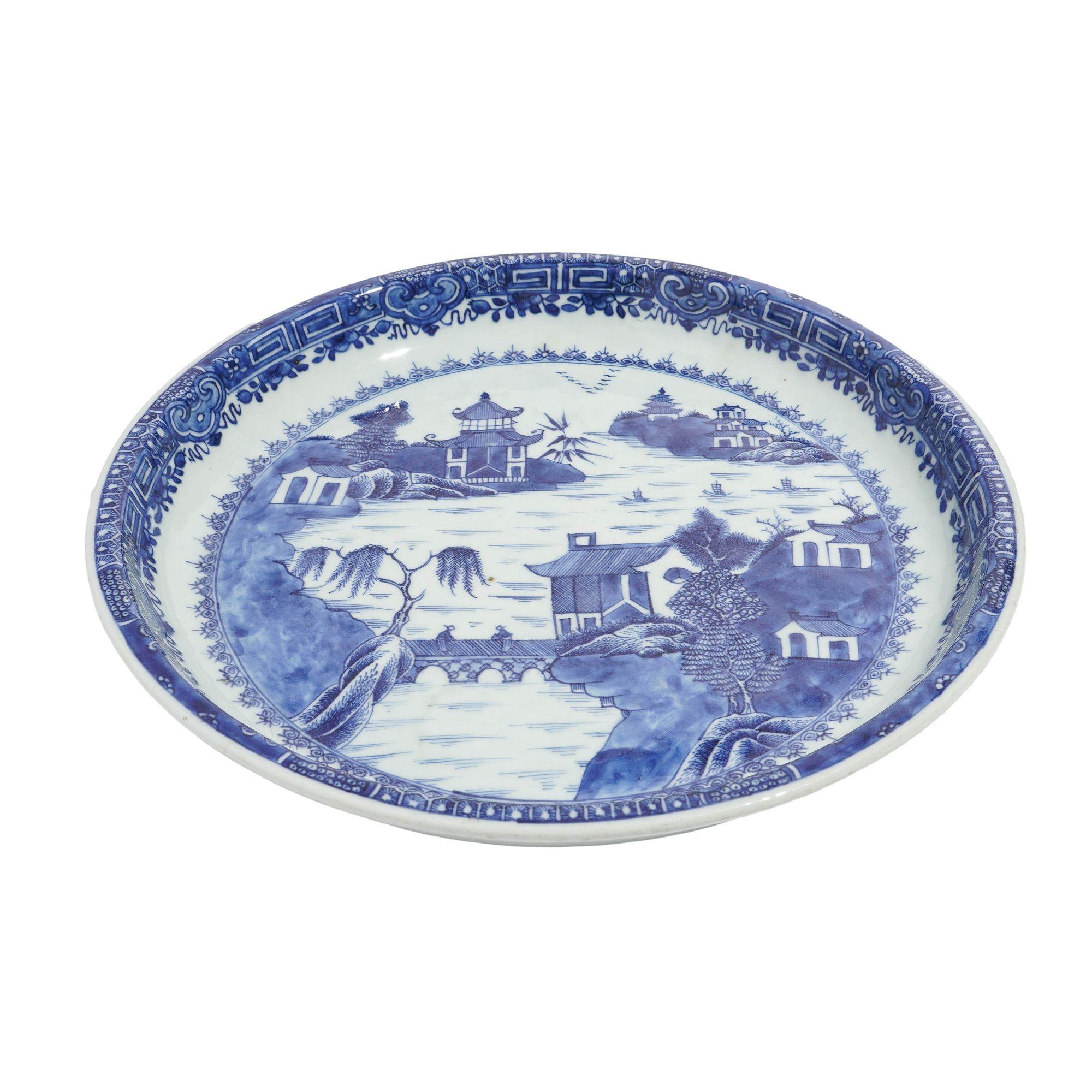 Chinese export porcelain charger hand decorated in cobalt underglaze blue centering on the Willow Tea House landscape. The deep standing rim is detailed with a classic Chinese running pattern. The foot of the charger is unglazed.

China, early