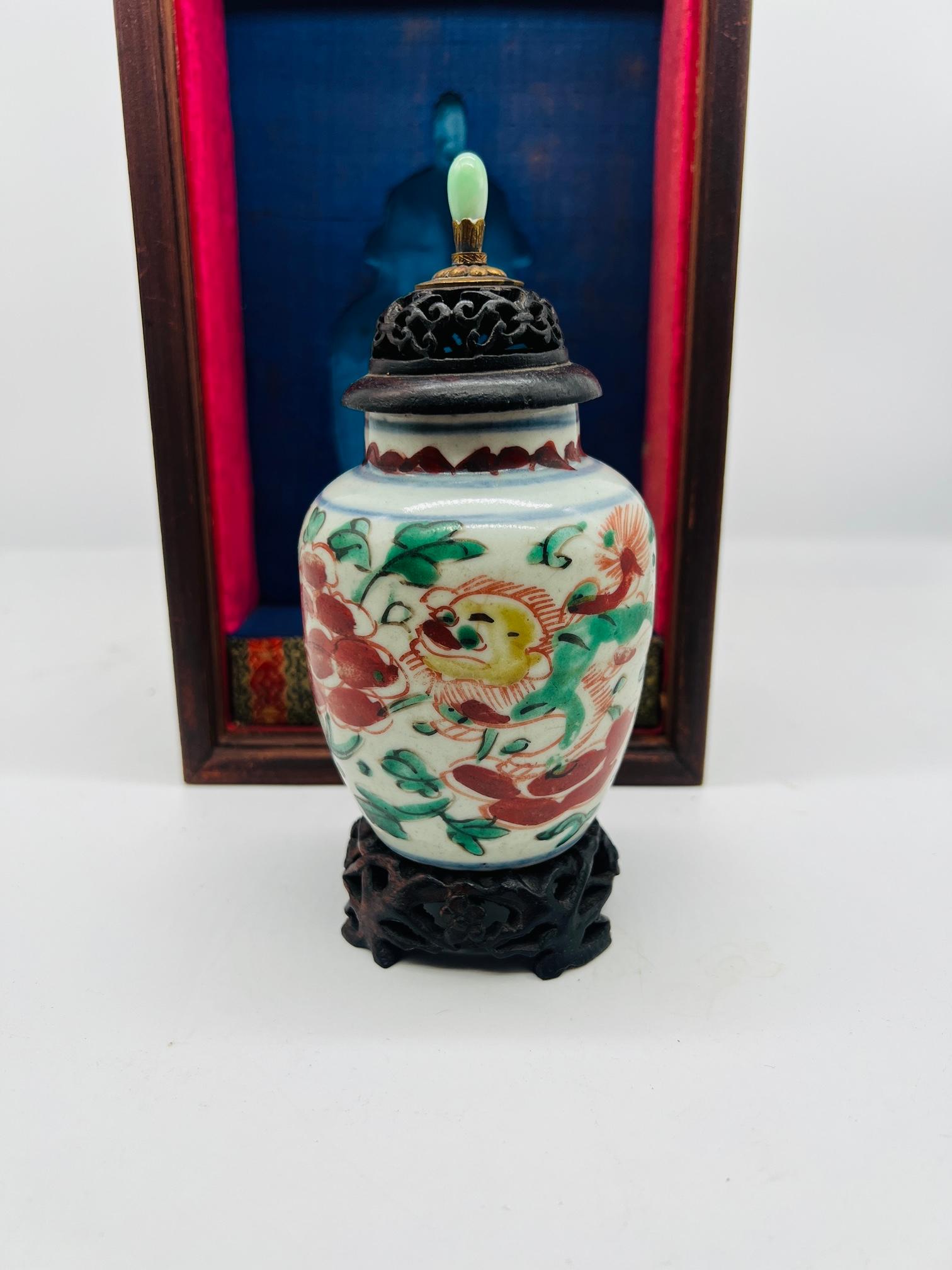 Chinese, 19th century or earlier.

An early Chinese porcelain vase polychrome decorated with a green, red and blue foo dog surrounded by foliate detailing. Accompanied with a rosewood stand, pierced rosewood lid mounted with a bronze and jade