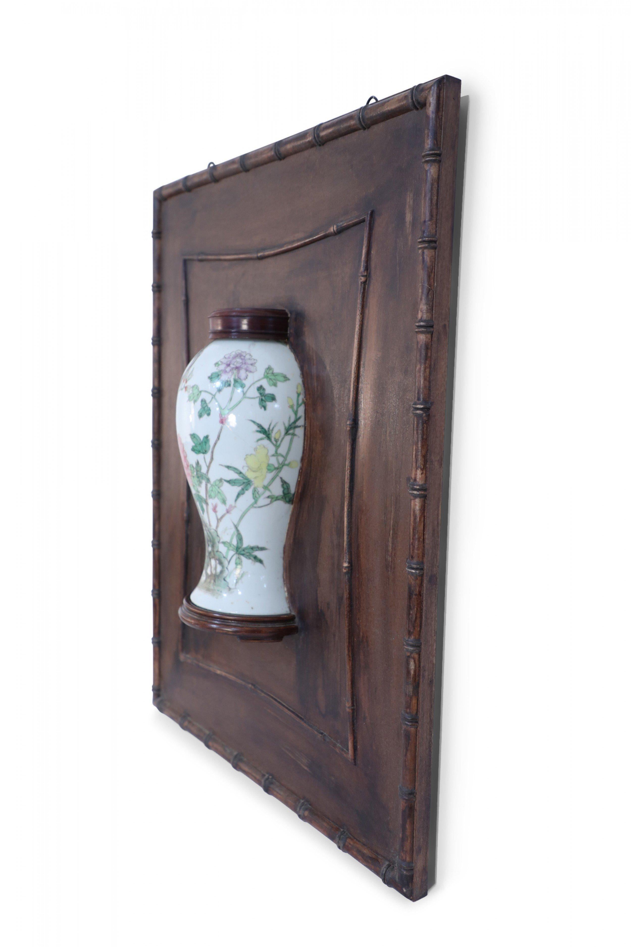 Chinese wall plaque framing a bisected famille rose porcelain lidded vase, depicting trees and blooming florals, in wood with a decorative faux bamboo edge.