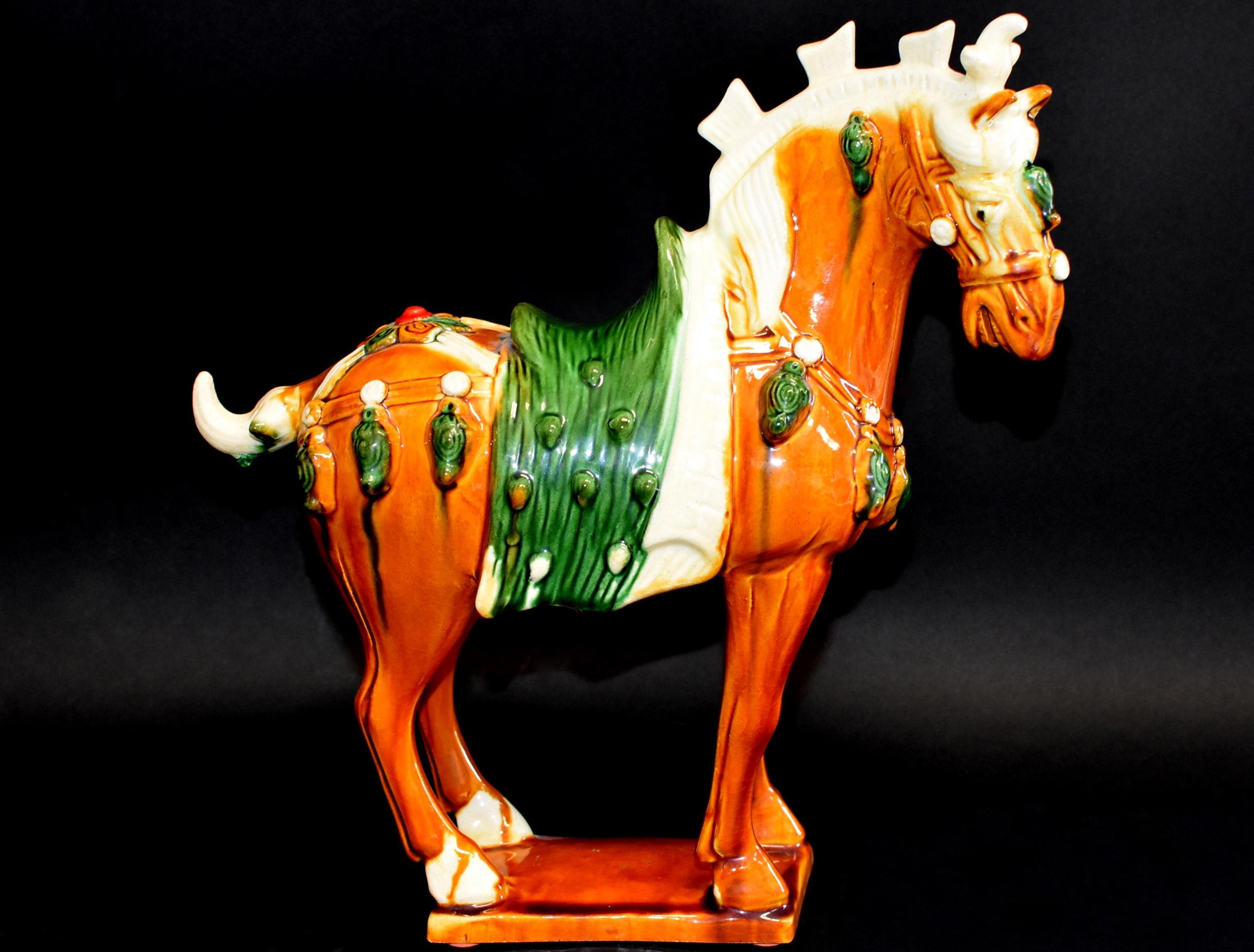 The horse is shown standing on a slab base with ears pricked and head turned slightly to the right, and is covered with a caramel glaze, the mane and tail with cream glaze. Both saddle and the harness, the latter hung with bells, are green glazed.