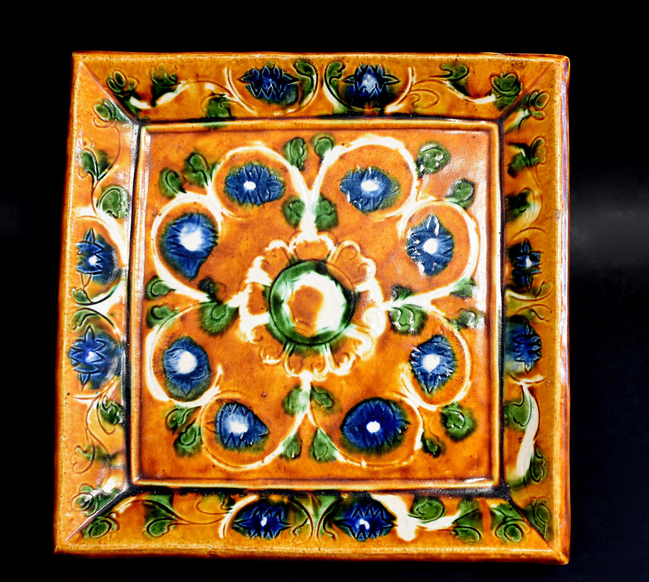 The square plate is beautifully painted in the center with a large bloom of white chrysanthemum, featuring a blue lotus on each petal, within a stylized design of radiating green buds on a caramel glazed ground below a rim decorated by repeating the