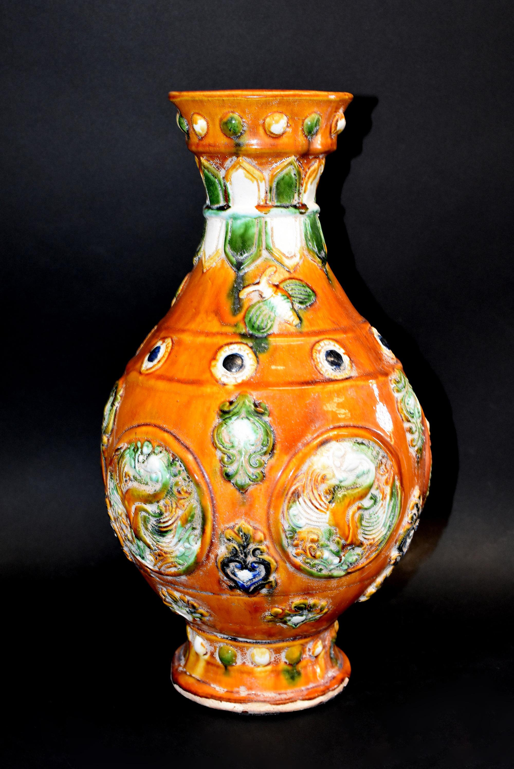 A Tang style sancai glazed pottery globular vase. The rounded body is applied with five rosettes, each featuring a flying phoenix, alternating with pairs of curved shields. The neck is decorated by a continuous pattern of double lotus petals above a