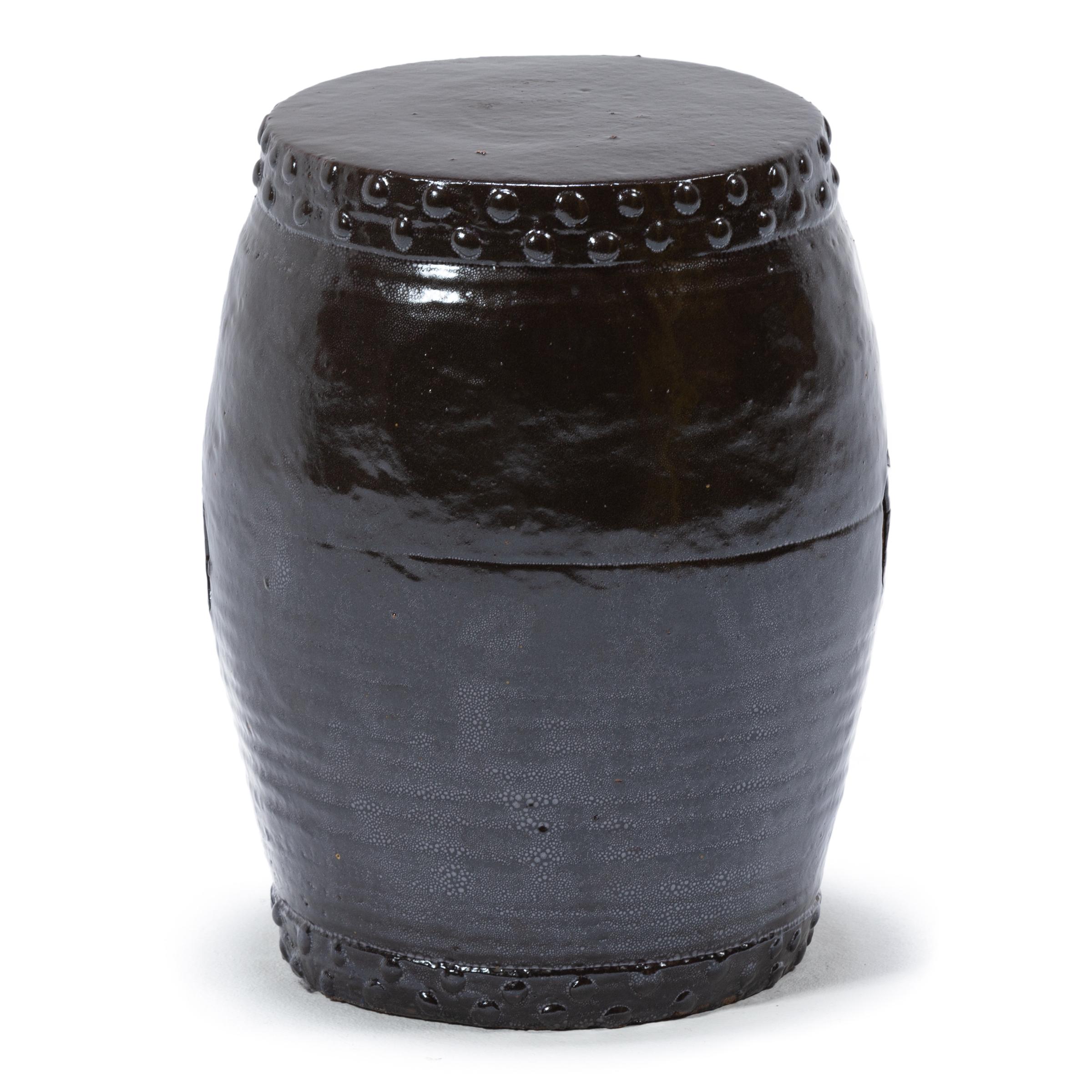 This early 20th century garden stool from China's Zhejiang province keeps with tradition with its simple drum-form shape, cloaked in a dark brown glaze. Ringing both the top and bottom, a pattern of boss-head nails imitates those used to stretch a