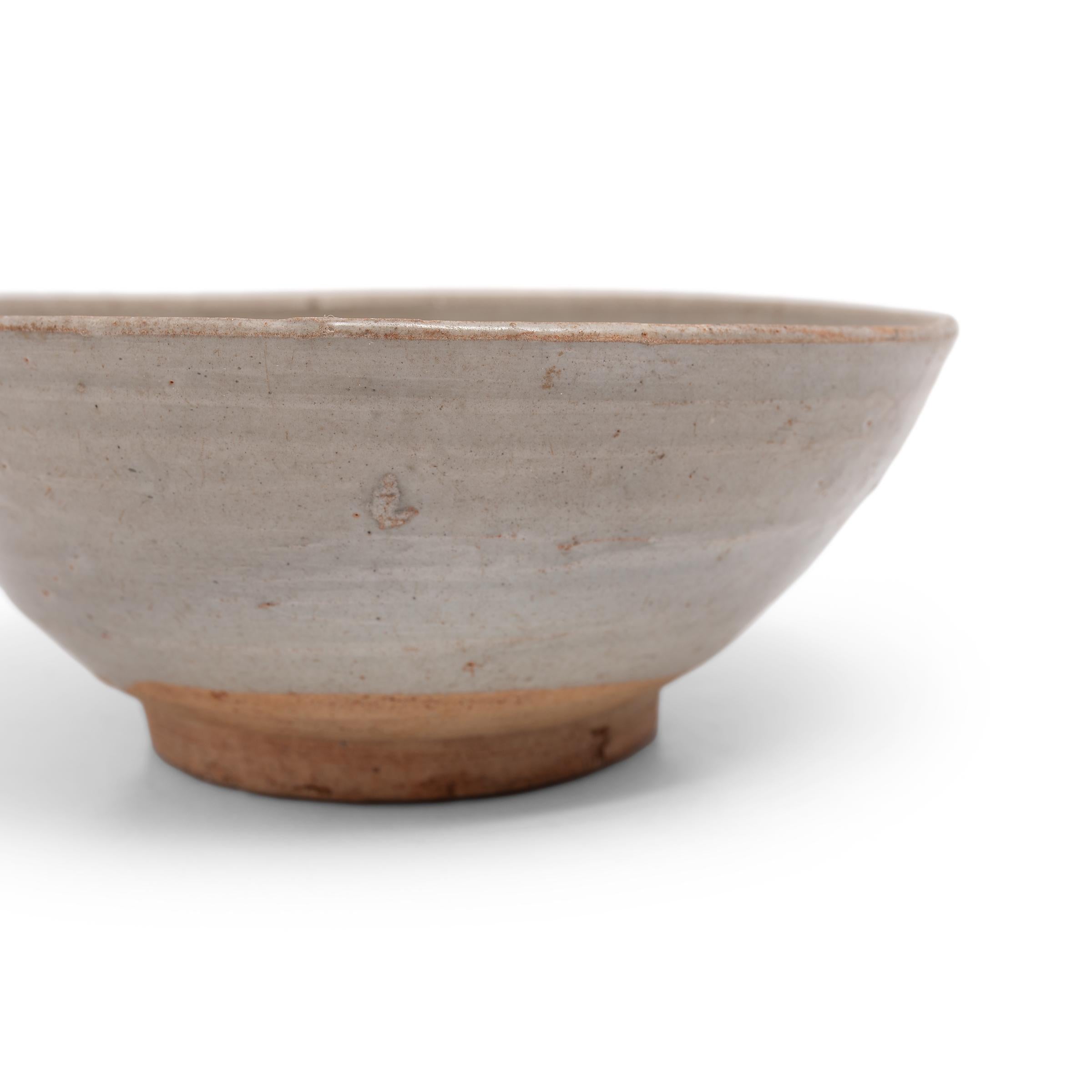 This 19th-century ceramic bowl is simply shaped with tapered sides and a heavy footed base. A cool grey glaze coats the bowl inside and out, pooling at the bottom with increased opacity. The footed base has been left unglazed, along with a circular