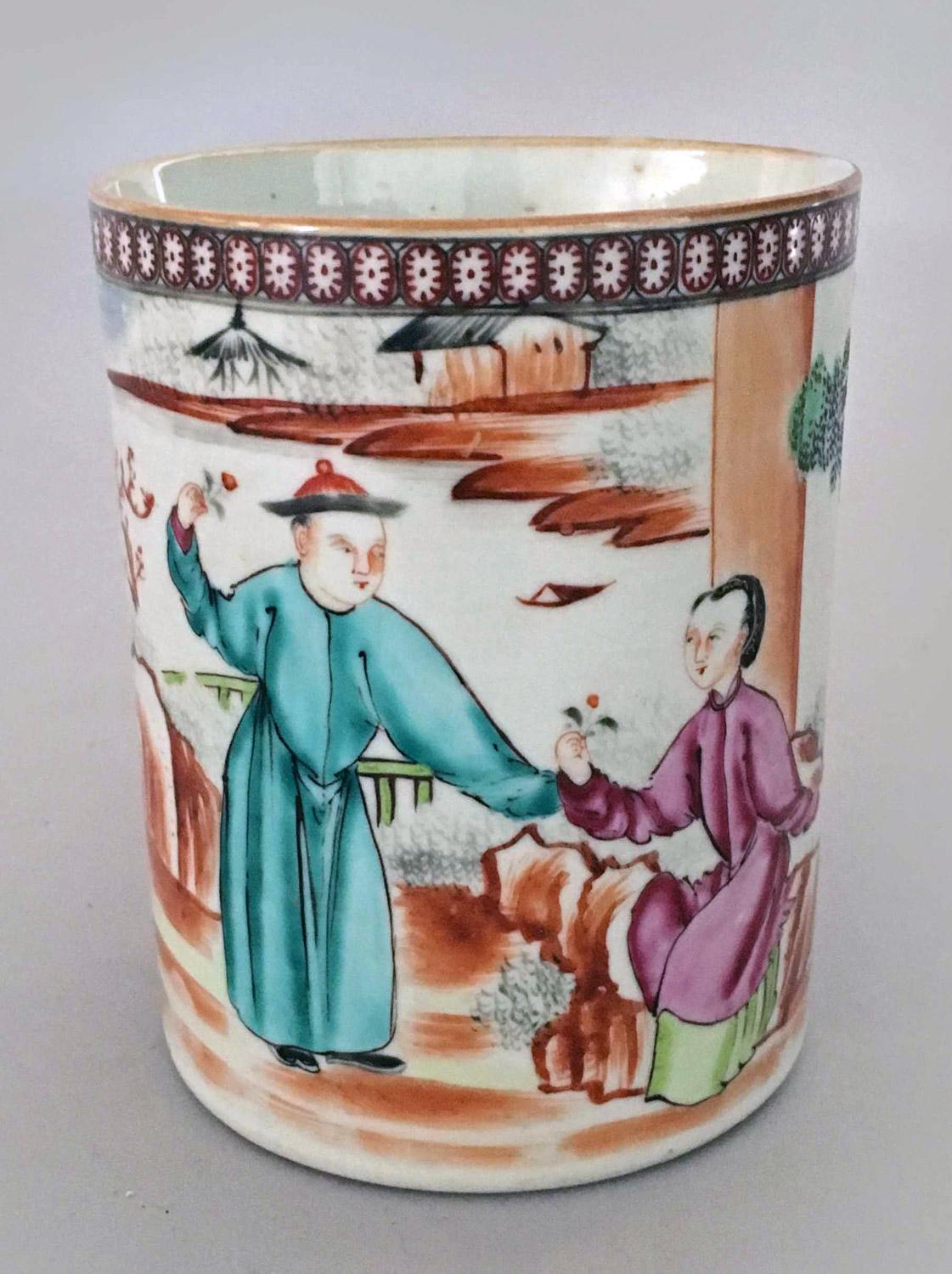 18th century Chinese Qianlong period porcelain famille rose mug depicting four figures, trees, rockery and a diaper border at the top edge of oval patterns. A turquoise-robed gentleman is presenting his lady clothed in pink and green with flowers.