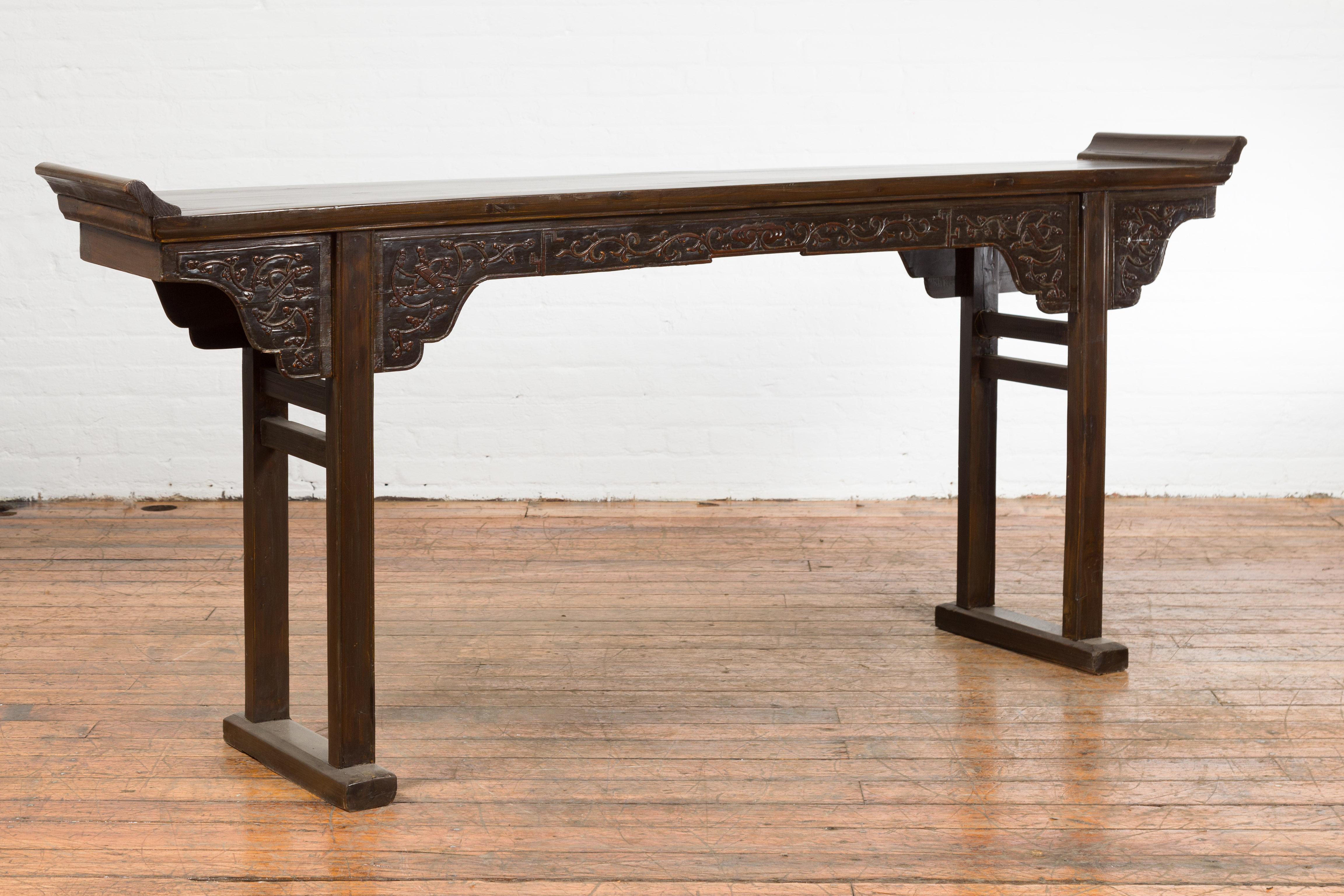 A Chinese Qing Dynasty period altar console table from the 19th century, with foliage-carved apron and everted flanges. Created in China during the Qing Dynasty period, this wooden altar console table features a long and narrow rectangular top