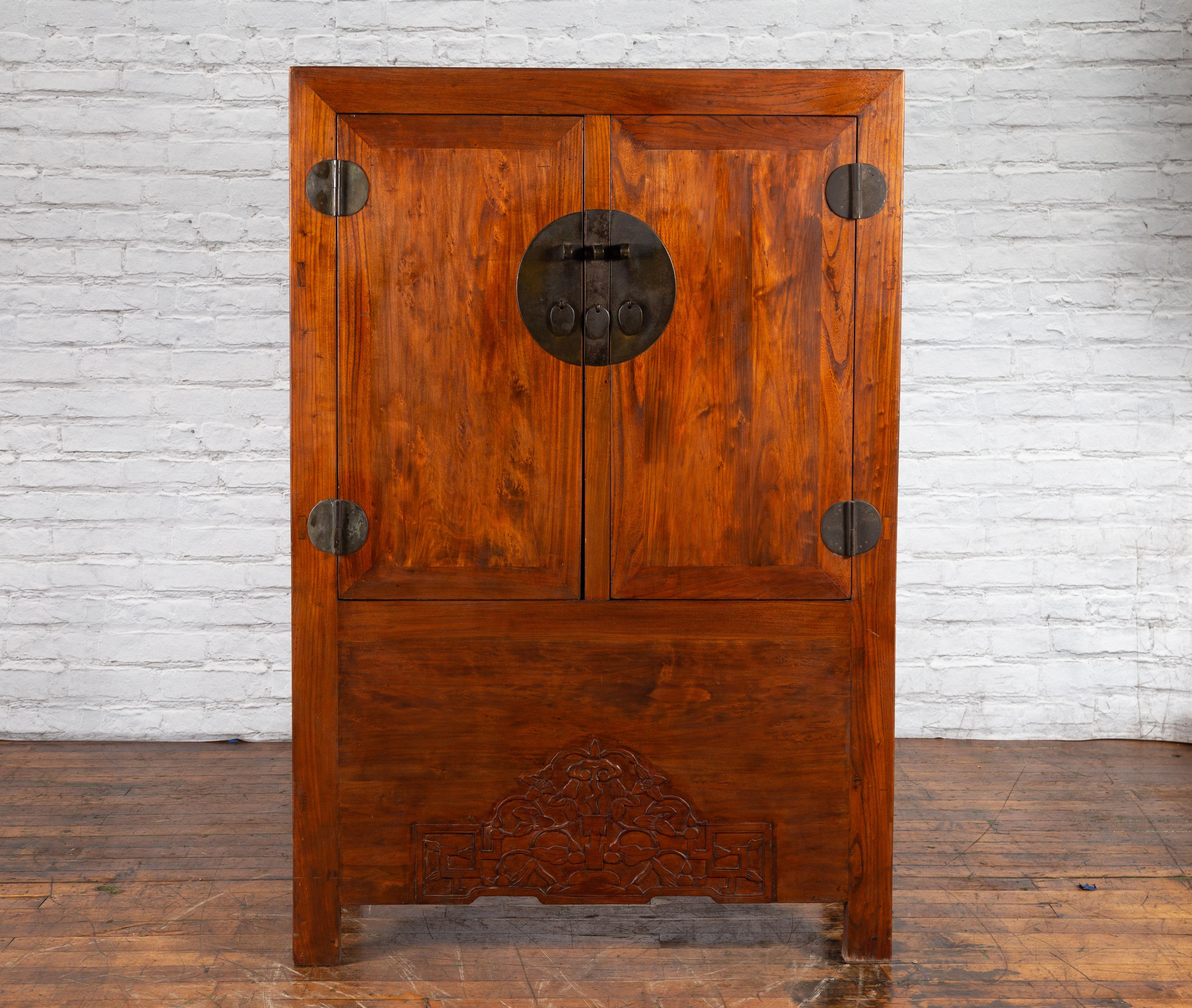 A Chinese Qing Dynasty period armoire from the 19th century with natural wood finish, carved skirt, hidden drawers and medallion hardware. Created in China during the Qing Dynasty period in the 19th century, this wooden armoire features a linear